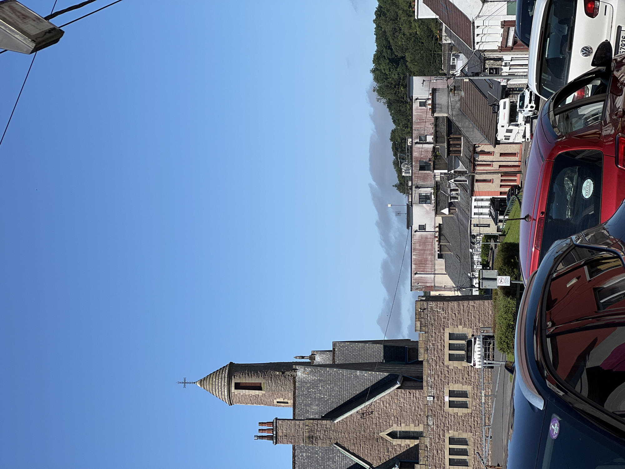 The image shows a street view in the town of Donegal, Ireland, on a sunny day with a clear blue sky. In the foreground, the tops of several cars are visible, parked along the street. To the left, a large stone building with a pointed tower and a cross on top, likely a church, dominates the scene. The building features slate roofs and arched windows. Behind the church, a row of townhouses and other buildings of varying heights and architectural styles line the street. Trees and a hill covered in dense green foliage can be seen in the background. The overall impression is of a typical Irish town on a pleasant day.