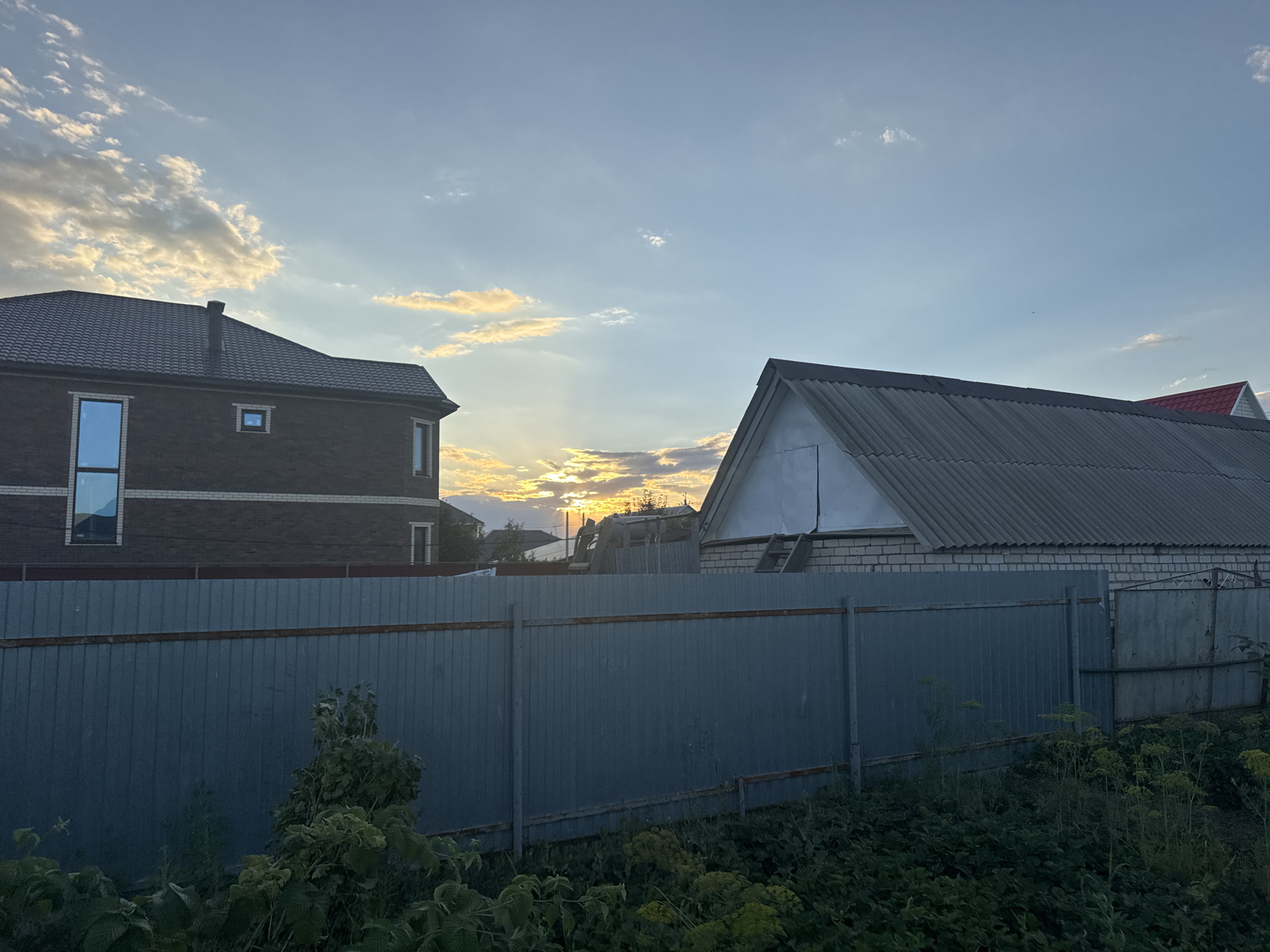 This image captures a suburban scene at dusk, likely in Zaşağan, Kazakhstan. The foreground is dominated by a tall, grey corrugated metal fence that stretches across the frame, partially obscuring the view of the properties behind it. Lush green vegetation, including tall weeds and leafy plants, grows densely in front of and beyond the fence, suggesting an unkempt or naturalized area.
Behind the fence, two buildings are visible. On the left, a dark brick house with a pitched, dark grey roof and a prominent blue-tinted window is seen. The house has a chimney and appears to be two stories tall. To the right of this house is another structure with a light-colored brick lower wall and a steeply pitched, dark grey corrugated metal roof. This building has a white gable end with a visible ladder leaning against it. Further to the right, the edge of another building with a red roof is partially visible.
The sky above is a gradient of pale blue, transitioning to warmer tones near the horizon where the sun is setting. Scattered clouds, tinged with orange and yellow from the setting sun, are visible against the blue. The sunlight is diffused, casting a soft light on the scene. The overall impression is one of a quiet residential area at the end of the day.