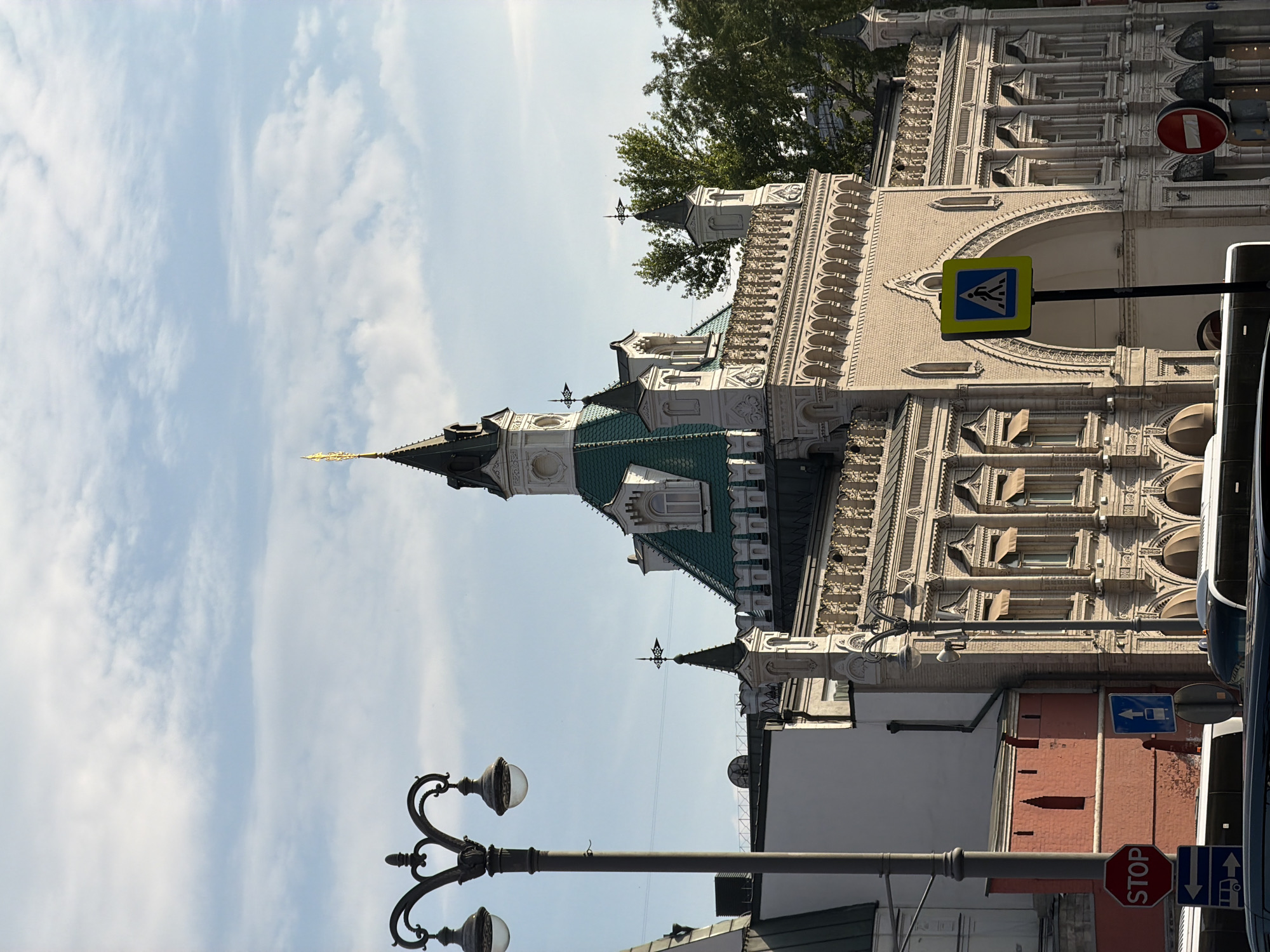 The image shows a street view in Moscow, Russia. The building is the GUM department store, with ornate facade and a green roof. A street lamp is visible in the foreground, and traffic signs are also present, including a pedestrian crossing sign and a STOP sign. The sky is visible with scattered clouds.