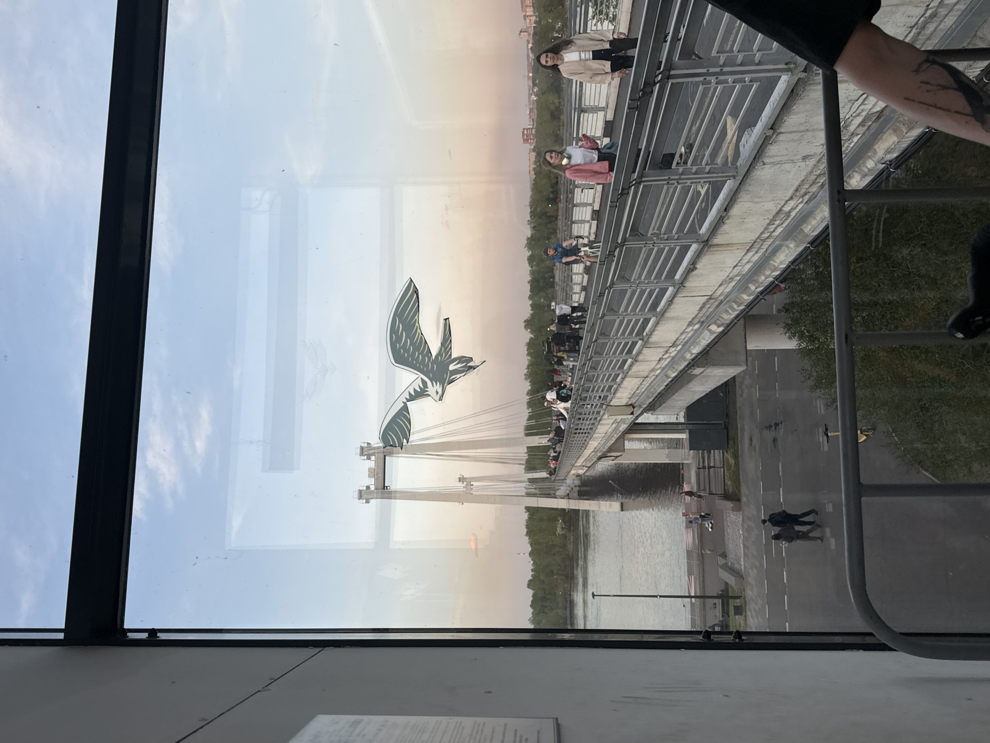 The image depicts a view from inside a building or structure looking out through a window at a bridge in Krasnoyarsk, Russia. The window frame is visible on the sides, and a reflection of the interior is partially visible in the glass.
The bridge is a pedestrian or mixed-use bridge with a suspension structure supported by cables and towers. There are stylized bird sculptures, seemingly seagulls, adorning the bridge's towers. People are seen walking, biking, and standing on the bridge.
Below the bridge is a body of water, possibly the Yenisei River, which flows through Krasnoyarsk. There is a road running along the riverbank with pedestrians walking on it. The overall environment suggests an urban setting during a time of day with soft, possibly evening light.