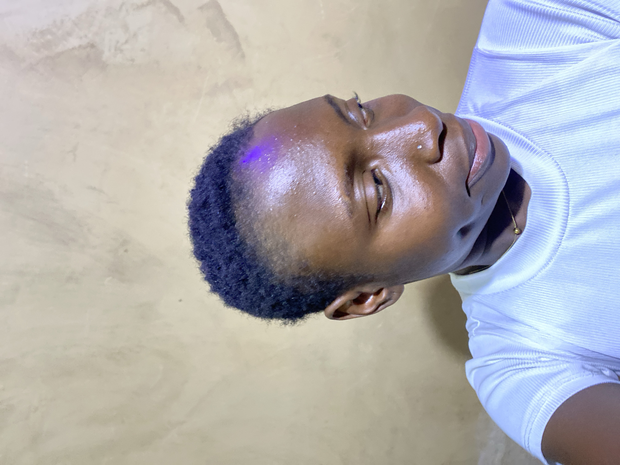 This close-up portrait, possibly taken in Kuru Dungus, Nigeria, features a person with dark skin and short, dark, curly hair. The individual is positioned with their head tilted slightly back and looking upwards, with their eyes closed or cast downwards, giving a contemplative or serene expression. A small silver piercing is visible on the left nostril.
The person is wearing a white, ribbed long-sleeved top, and a thin gold chain can be seen around their neck. Their skin exhibits a glossy sheen, particularly across the forehead and cheeks, which might be due to natural oils, sweat, or a deliberate highlight. A distinct, subtle purple light or reflection is visible on the left side of their forehead and hairline.
The background is a plain, light beige or tan surface, likely a wall, with a slightly textured and uneven appearance. The lighting is bright, creating strong highlights on the subject's skin and clothing. There is no visible text in the image, and no specific activity is taking place beyond posing for the photograph. The time of day is not discernible from the image, but the lighting suggests an indoor setting or controlled light source.