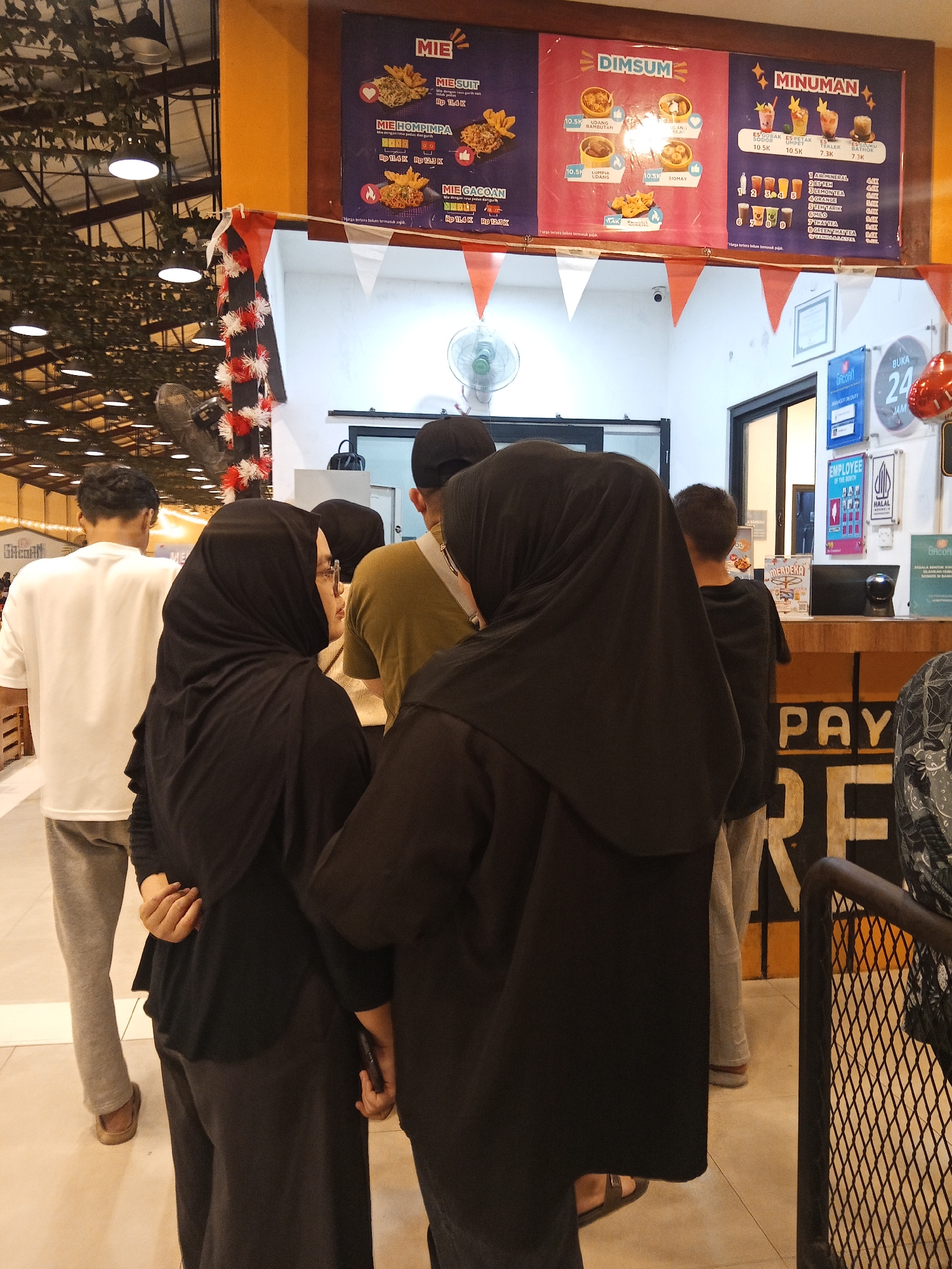 In Lubuklinggau, Indonesia, a group of individuals stand in line at a food stall with a menu board displaying "MIE," "DIMSUM," and "MINUMAN." The menu board is colorful and features images of food and drinks with their prices. Several people are visible, including a person in a white t-shirt and grey pants, two individuals wearing black hijabs, one person in a light brown t-shirt, and another in a dark t-shirt and grey pants. The environment appears to be an indoor market or food court, with a decorative ceiling, lighting fixtures, and a fan. A counter with a cash register and various signs are also present. The overall scene suggests a casual dining or takeout experience.