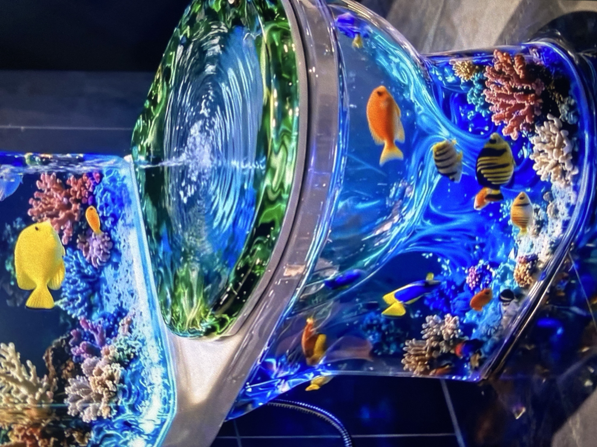 The image shows a toilet-shaped aquarium, filled with water, fish, and coral. The 'toilet bowl' is filled with blue-tinted water. The 'toilet seat' is made of clear material with a green-tinted ring. Various fish, including yellow, orange, and black striped ones, swim among coral structures. A flexible silver hose is visible at the bottom of the image, near the base of the aquarium. The setting appears to be indoors. The city of Lhokseumawe, Indonesia is not discernible from the image.
