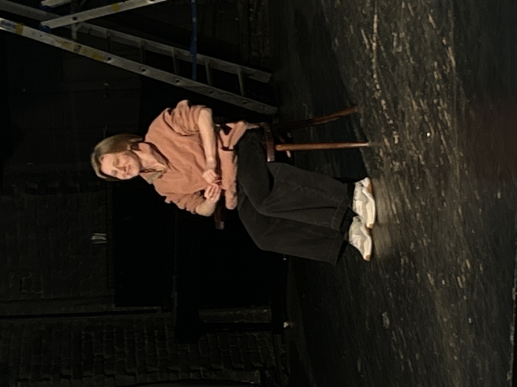 The image depicts a single person seated on a wooden chair in a dimly lit, dark environment, likely an indoor space such as a theater stage, backstage area, or rehearsal room. The person has short, light brown hair and is wearing a loose-fitting, long-sleeved top in a salmon or dusty pink color, dark wide-legged pants, and white sneakers. They are leaning forward slightly, with their arms crossed over their lap, and are looking towards the viewer with a gentle smile or thoughtful expression, appearing relaxed.
The background is predominantly dark. To the upper left, a metal ladder or part of a scaffolding structure is visible. The floor and a section of what appears to be a wall on the right are dark and have a rough, textured appearance, possibly aged wood, concrete, or a painted backdrop. The lighting is localized, highlighting the person while keeping the surroundings in deep shadow, suggesting artificial stage or studio lighting. The setting conveys a utilitarian and functional space. The time of day is not discernible. No text is visible in the image.