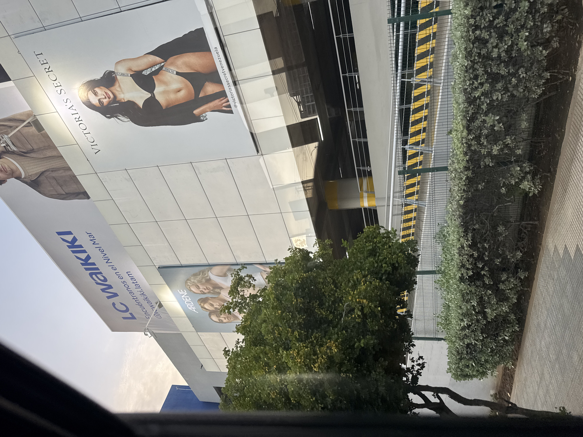 The image shows a large commercial building in Porlamar, Venezuela, featuring several prominent billboards. The largest billboard displays a woman in lingerie and the text "VICTORIA'S SECRET." Below this, another billboard shows three women with the brand name "ARDENE." Further to the left, a billboard for "LC WAIKIKI" is visible, with the text "Encuéntranos en el Nivel Mar" and "@lcwaikiki.latam." The building appears to be a mall or shopping center, with multiple levels of parking visible behind a railing. In the foreground, a well-maintained hedge and a small tree line a paved walkway. The sky is overcast, suggesting it is either early morning or late afternoon. The overall scene depicts a retail environment in an urban setting.