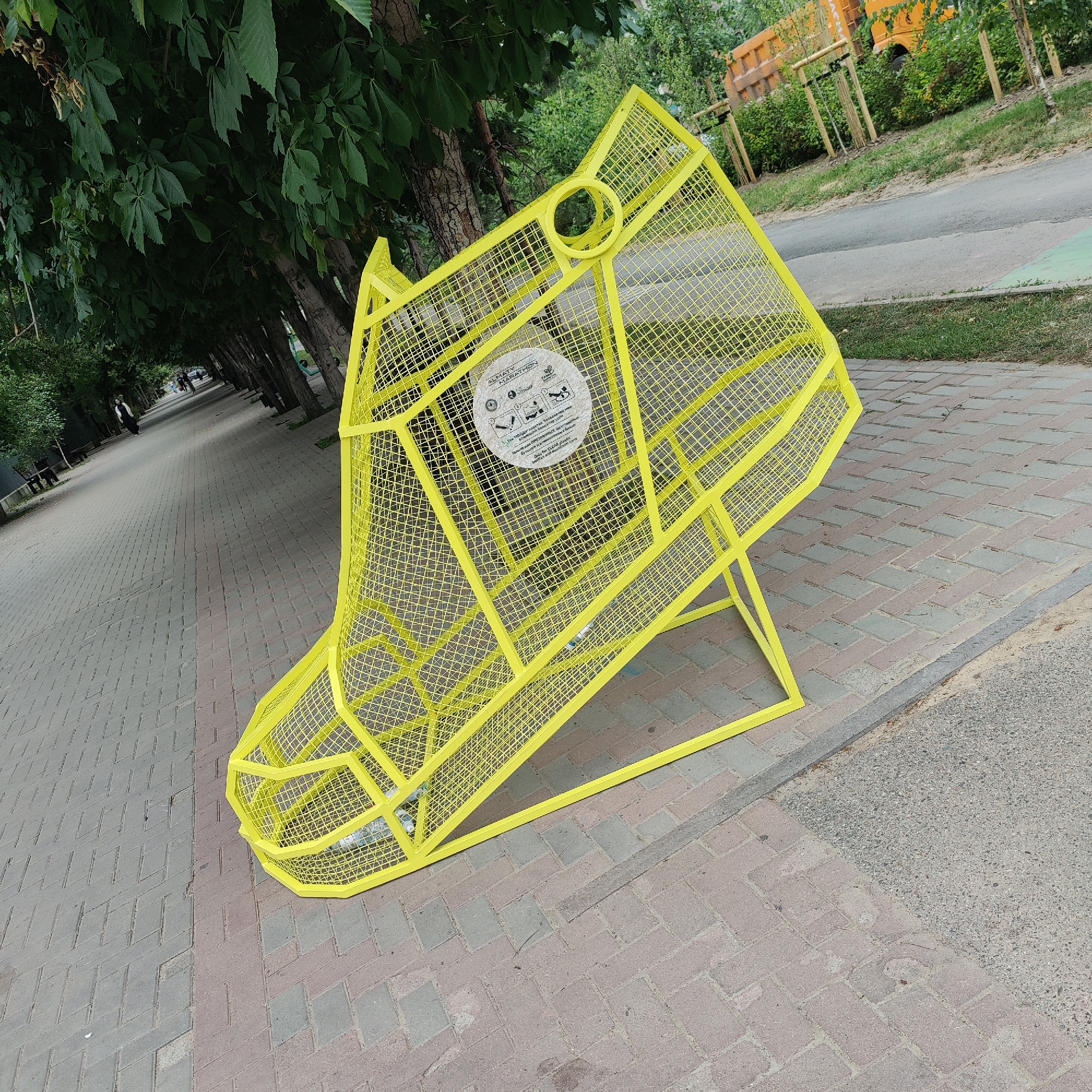 An outdoor scene depicts a bright yellow, mesh trash receptacle shaped like a dog's head, situated on a paved pedestrian walkway. The receptacle has a circular opening at the top, resembling an ear, and a larger opening at the front for depositing waste. A circular sticker with text and graphics is affixed to the side of the receptacle. The walkway is bordered by a strip of grass and a road. In the background, trees line the walkway, casting shadows on the path. Beyond the trees, a section of a city street is visible, with buildings and a yellow truck in the distance. The scene is set during daylight hours, with overcast sky conditions. The image is taken from a slightly elevated angle, looking down towards the walkway and receptacle. The overall atmosphere is urban and public, suggesting a park or street setting.