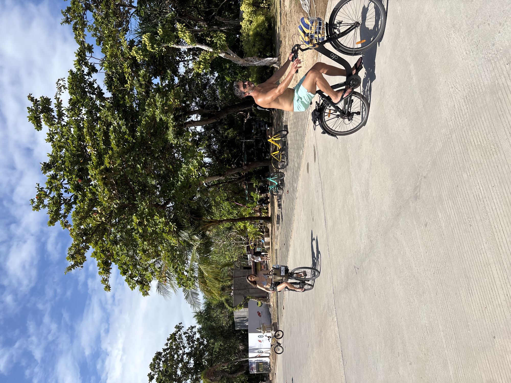 In Kuta Selatan, Indonesia, a man and a woman are cycling on a paved road bordered by lush green trees and vegetation under a bright blue sky with scattered white clouds. The man, with a beard and sunglasses, is shirtless and wearing light blue swim shorts, riding a black bicycle with a basket containing a yellow and blue striped towel. The woman, in a patterned bikini top and dark shorts, is on a black bicycle with a basket and a bag, also wearing sunglasses. In the background, several other bicycles, including yellow and blue ones, are parked or in motion, and there are indications of construction or development with concrete structures and fencing visible. The scene suggests a leisurely outdoor activity in a tropical or subtropical environment.