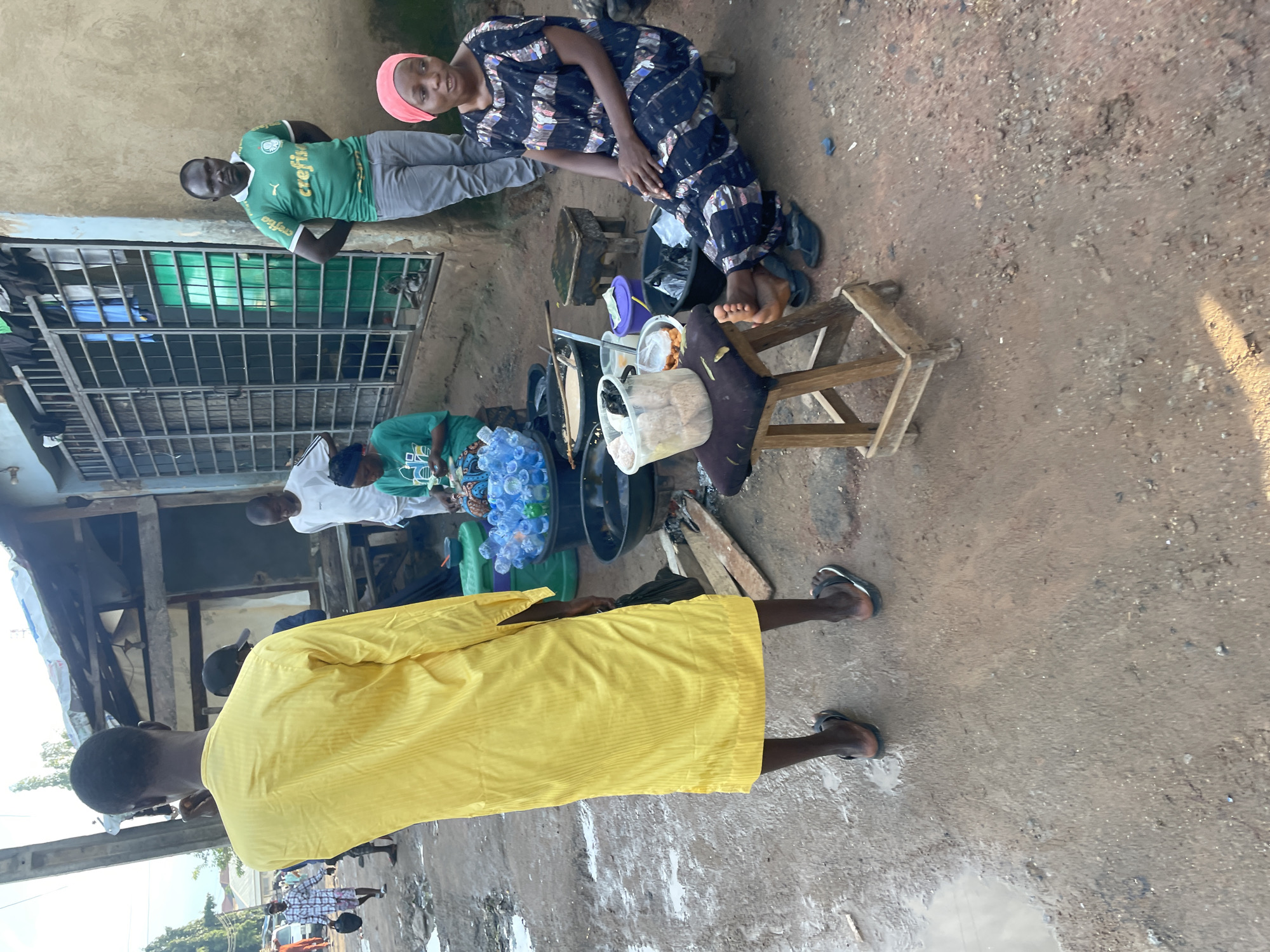 This image depicts a street scene in Abuja, Nigeria, likely in a bustling commercial area. In the foreground, a man wearing a bright yellow tunic stands with his back to the camera. He is facing a food stall where a woman in a blue and white patterned dress and a pink headscarf is seated on a low stool, attending to customers or preparing food. She is looking to her right. To her left, another woman in a green shirt is seated behind a display of bottled water. A man in a green "Crefisa" t-shirt and grey trousers stands to the right of the food stall, looking towards the camera. The ground is dusty and uneven, with puddles of water scattered about, suggesting recent rain or poor drainage. In the background, there are simple structures that appear to be shops or stalls, with metal bars on the windows. Other people can be seen in the distance, walking along the street. The overall impression is of a lively, everyday scene in a Nigerian city.