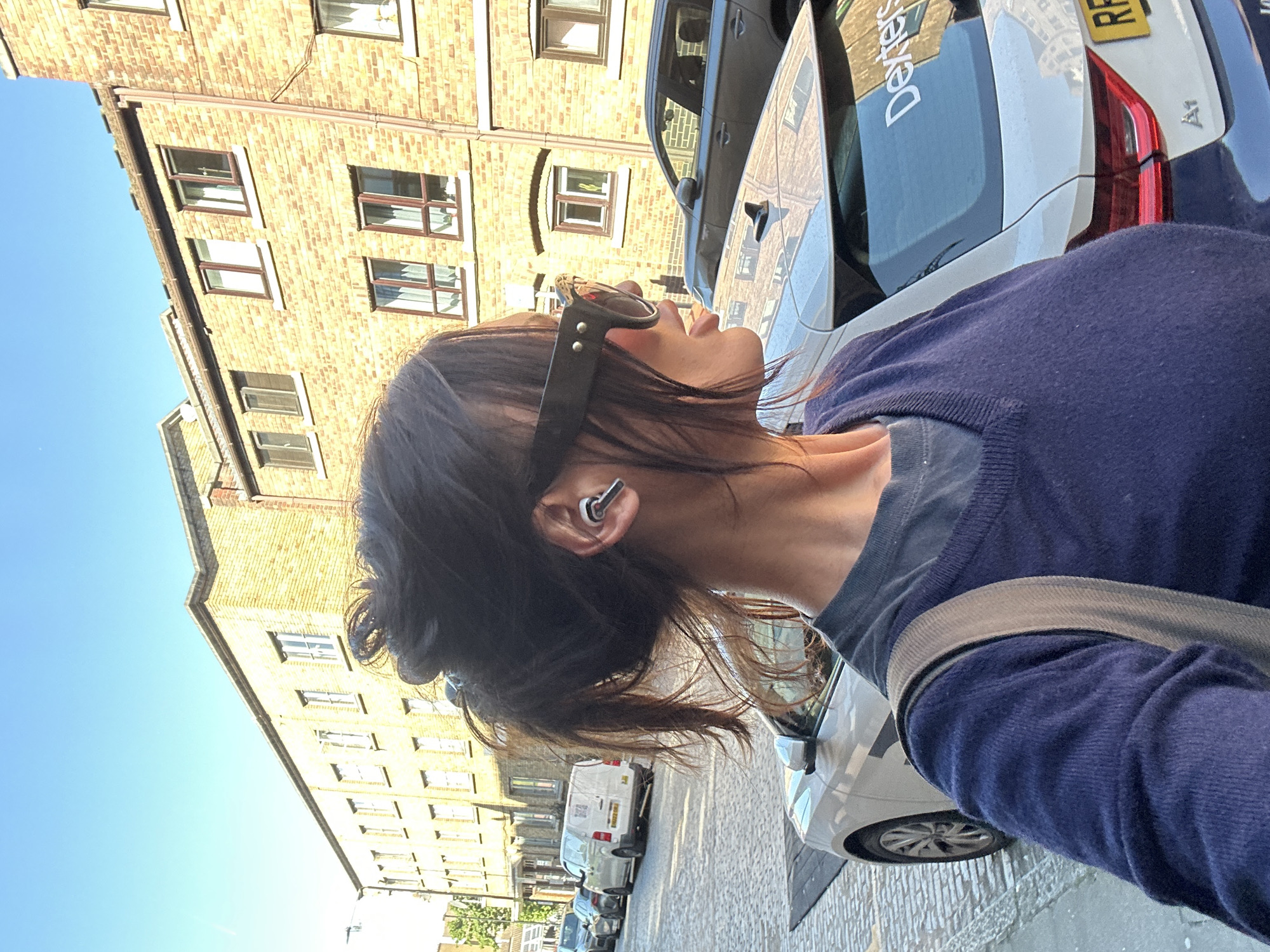 The image captures an outdoor urban street scene in London, United Kingdom, on a bright and sunny day. The primary subject is a person, seen from the right side of their head down to their upper torso, looking upwards and towards the left. They have dark hair, some of which is pulled back, with loose strands around their neck. Distinctive black sunglasses with silver studs are pushed up onto their forehead, and a white earbud is visible in their right ear. The person is wearing a navy blue long-sleeved top layered over a lighter blue garment, with a grey strap, likely from a bag, crossing their chest.
In the background, a street is lined with several parked cars and multi-story buildings constructed from light brown brick, a common architectural style in London. The buildings feature numerous windows with brown frames. The street itself appears to be paved with cobblestones. Among the parked vehicles, a white hatchback is prominent, displaying a "Dexters" logo on its rear window—a well-known London real estate agency. A partially visible yellow UK license plate, showing "RP," is also seen on this car. A dark grey car and a white van are also parked further down the street. The sky is a clear blue, and the strong sunlight casts distinct shadows, suggesting either morning or late afternoon.