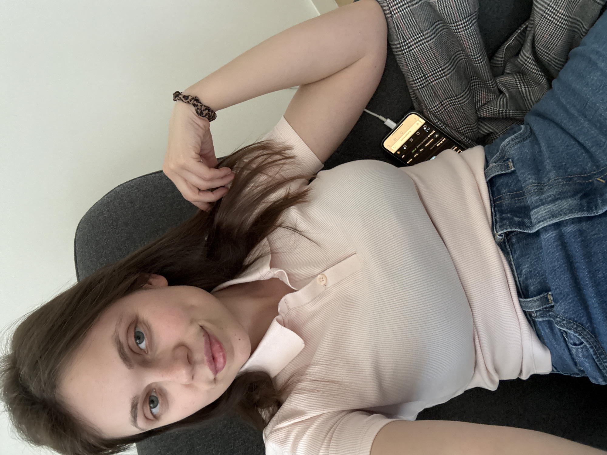 A woman lies reclined on a gray sofa. She wears a light pink polo shirt and blue jeans. Her hair is brown. She is looking at the camera. Her left hand touches her hair, and her right arm rests above her head. A phone charges next to her with a white cable. A gray and white plaid jacket lies next to the phone. The location appears to be an interior space. There is no clear indication of the city of Belgrade, Serbia.