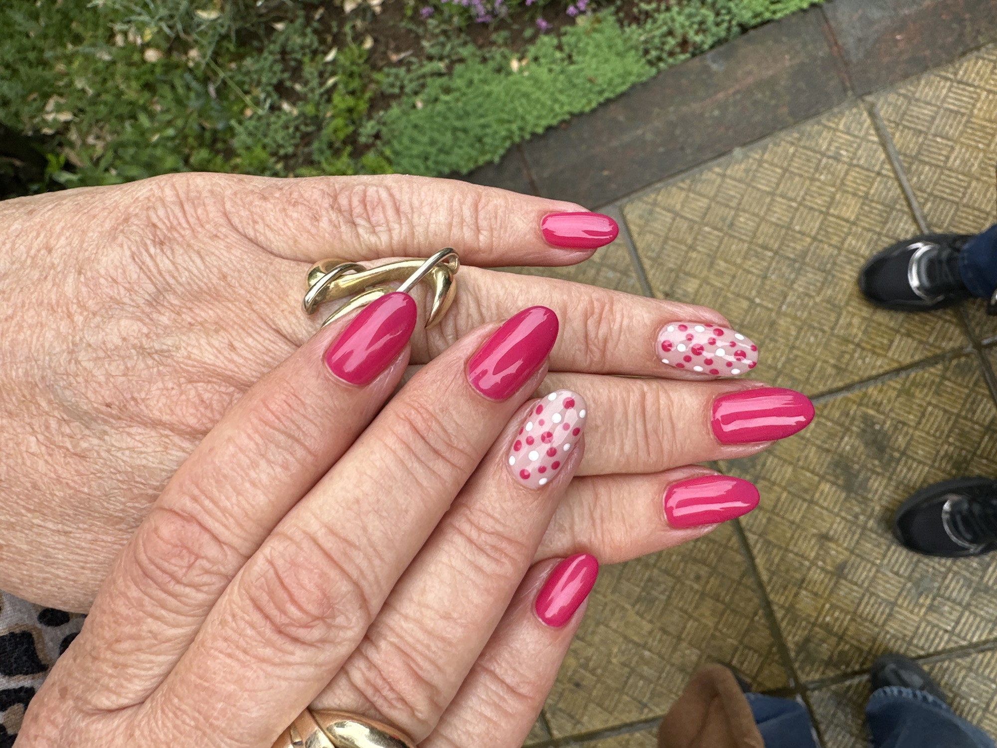 The image displays a pair of hands with manicured nails, primarily painted in a solid bright pink color, with two nails on one hand adorned with white and pink polka dots. The hands are positioned against a blurred background that appears to include outdoor elements such as greenery and a paved surface. The foreground features the hands prominently, with attention drawn to the nail polish and the texture of the skin. A gold ring with an intertwined design is worn on one of the fingers. The bottom of the frame captures the edge of the pavement and footwear of other people in the vicinity. The location is not specified within the visual details of the image.