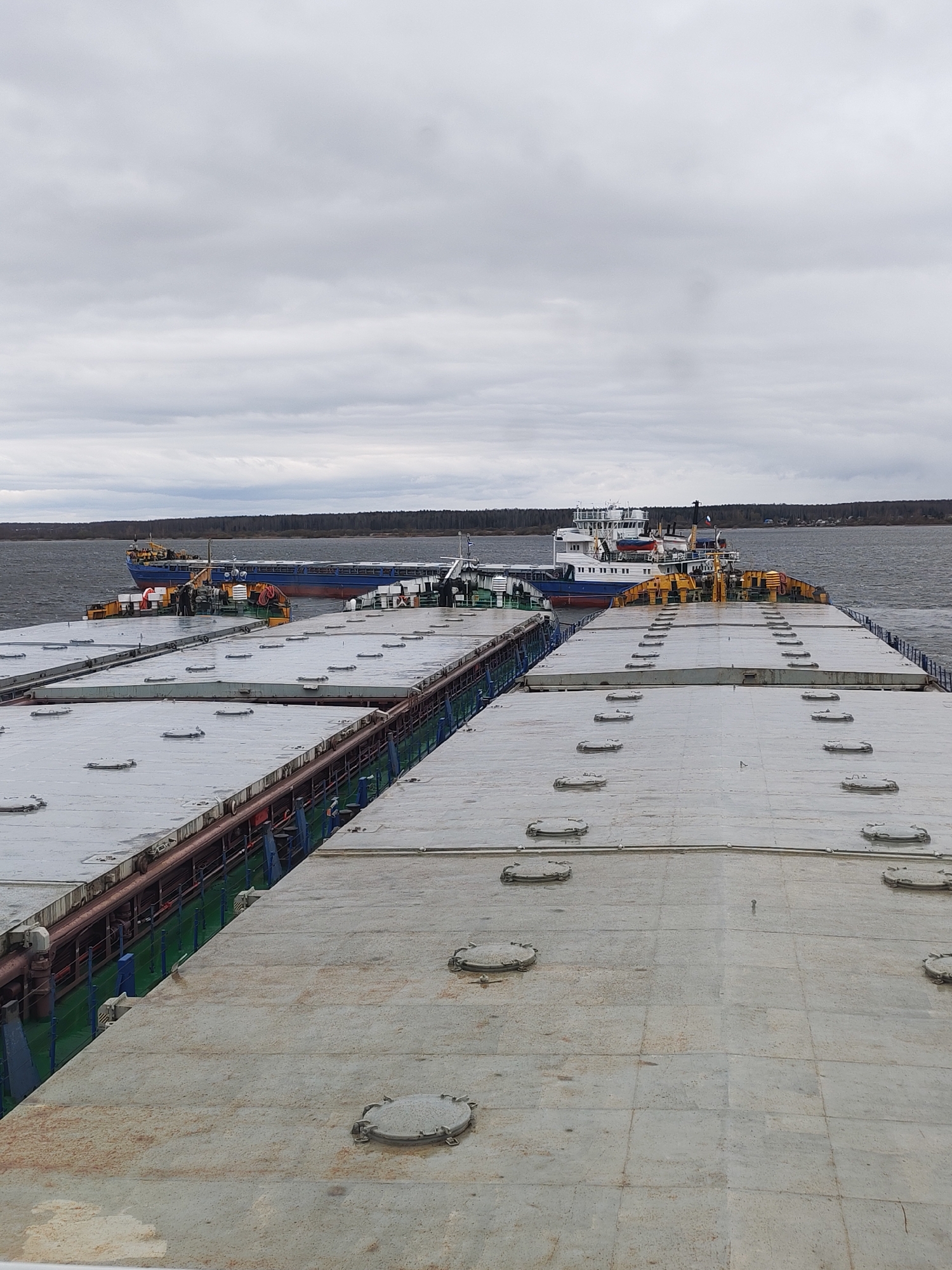 The image captures a view from the deck of a large industrial vessel, looking out across a wide body of water. In the immediate foreground, the grey, flat deck of the viewer's vessel is visible, covered with numerous circular, bolted hatch covers. Along the left side, exposed pipelines and railings are seen running parallel to the edge of the deck.
In the midground, two large vessels are prominent. To the left, a long, blue-hulled cargo ship or barge is visible, appearing to be coupled or sailing alongside the foreground vessel. Its deck is also flat and grey, suggesting it too is a cargo carrier. To the right, slightly behind and partially obscured, is another vessel, predominantly white with blue accents, possibly a tugboat or a service vessel, featuring a multi-story cabin structure and a visible red life raft on its deck. Both vessels appear to be either part of a convoy or are moored together on the water.
The background consists of a vast expanse of choppy grey water, leading to a distant, tree-lined shoreline that appears to have some scattered buildings or a small settlement. The sky above is entirely overcast with dull grey clouds, indicating a cloudy or stormy day. The overall scene suggests an industrial marine setting on a river or large lake in Bol'shie Mkhi, Russia, under muted, overcast conditions. There is no discernible text visible on any of the vessels or in the landscape.