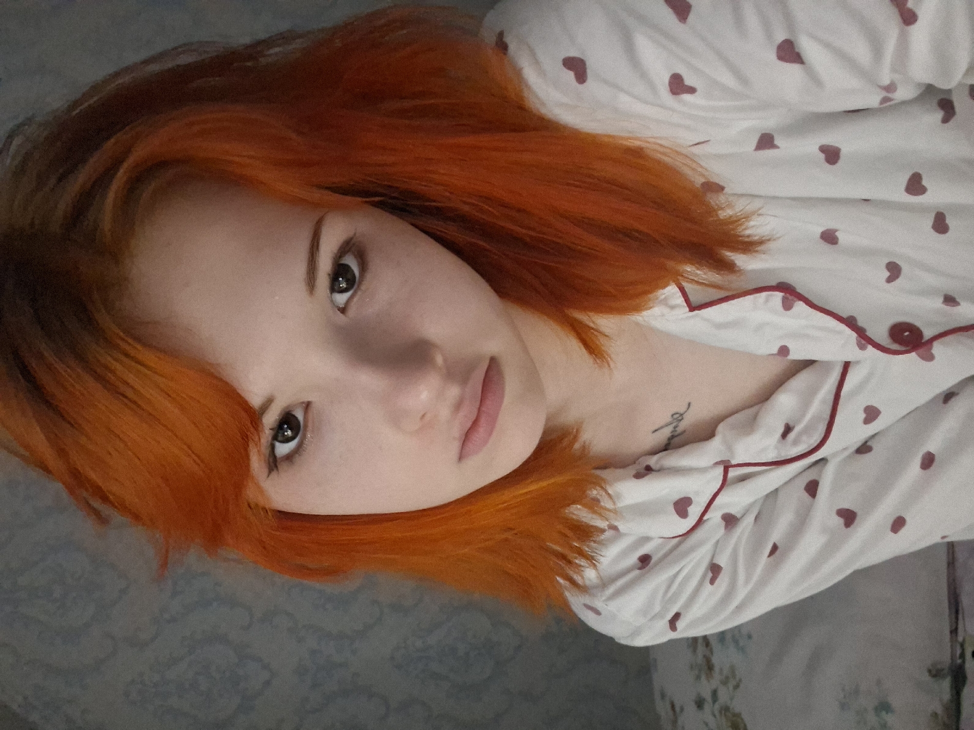 A young woman with bright orange hair is the focal point of a medium shot taken from a slightly elevated angle. She is wearing a white pajama top patterned with small, dark pink hearts and trimmed with dark pink piping. A small tattoo is visible on her chest. The background appears to be a bedroom or living space, with a patterned blanket or duvet visible. The location is Chaikovsky, Russia.