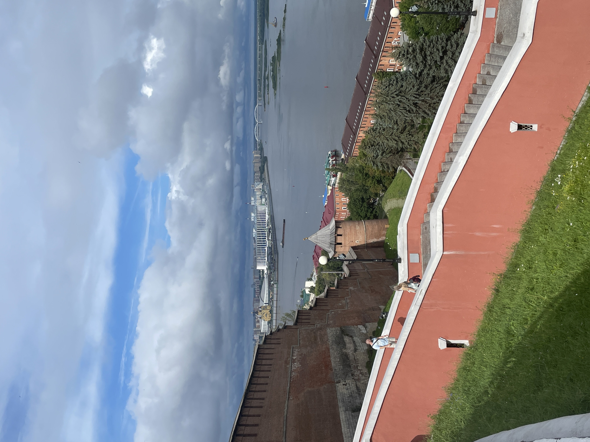 The image captures a panoramic view of Nizhny Novgorod, Russia, focusing on the Volga River and the historic Kremlin walls. In the foreground, a vibrant coral-colored staircase with white trim ascends, bordered by lush green grass. Two individuals are walking down the stairs: a man in a light blue t-shirt and shorts, and a woman in a white top and dark pants.
The middle ground features the imposing red brick walls of the Nizhny Novgorod Kremlin, with crenellated battlements and a distinctive round tower. Beyond the Kremlin walls, the wide expanse of the Volga River dominates the scene, dotted with a few boats and barges. In the distance, the city skyline is visible, characterized by modern buildings and bridges spanning the river. The sky is dramatic, with a mix of heavy, dark clouds and patches of bright blue. The overall impression is one of historical grandeur and natural beauty under a dynamic sky.