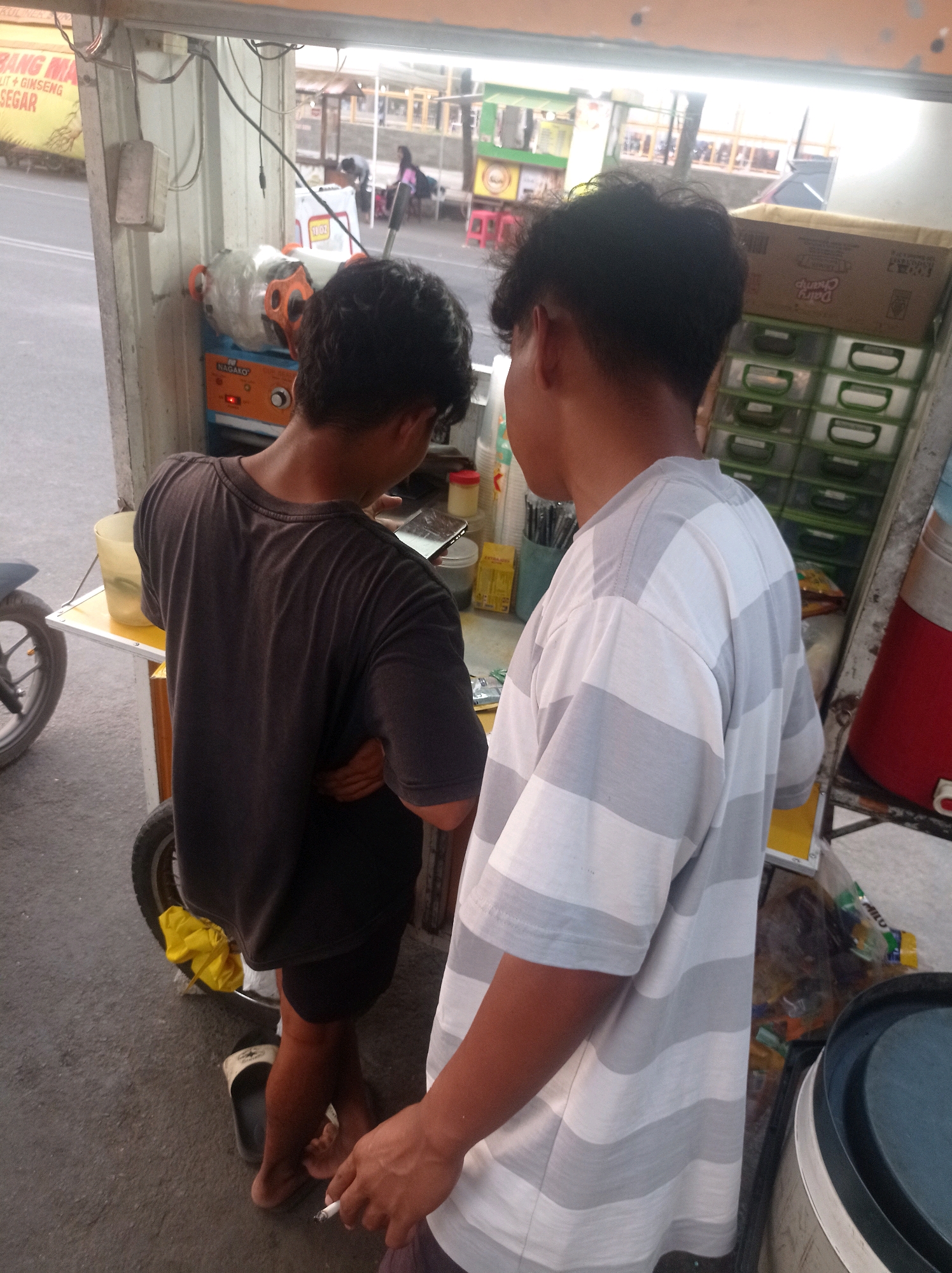In Tuban, Indonesia, two young men are standing near a food cart. The man on the left, wearing a dark t-shirt, is looking down at his phone, which is resting on a yellow surface. He is barefoot and wearing dark shorts. The man on the right, wearing a grey and white striped t-shirt, is holding a lit cigarette in his right hand. He is facing away from the camera, towards the food cart, which has several green drawers and a large red container. The food cart is situated on a paved surface, likely a street or sidewalk, with a grey motorcycle wheel visible on the left side of the frame. In the background, there are some buildings and other people, suggesting a public area. The lighting indicates it is daytime, possibly late afternoon or early evening.
