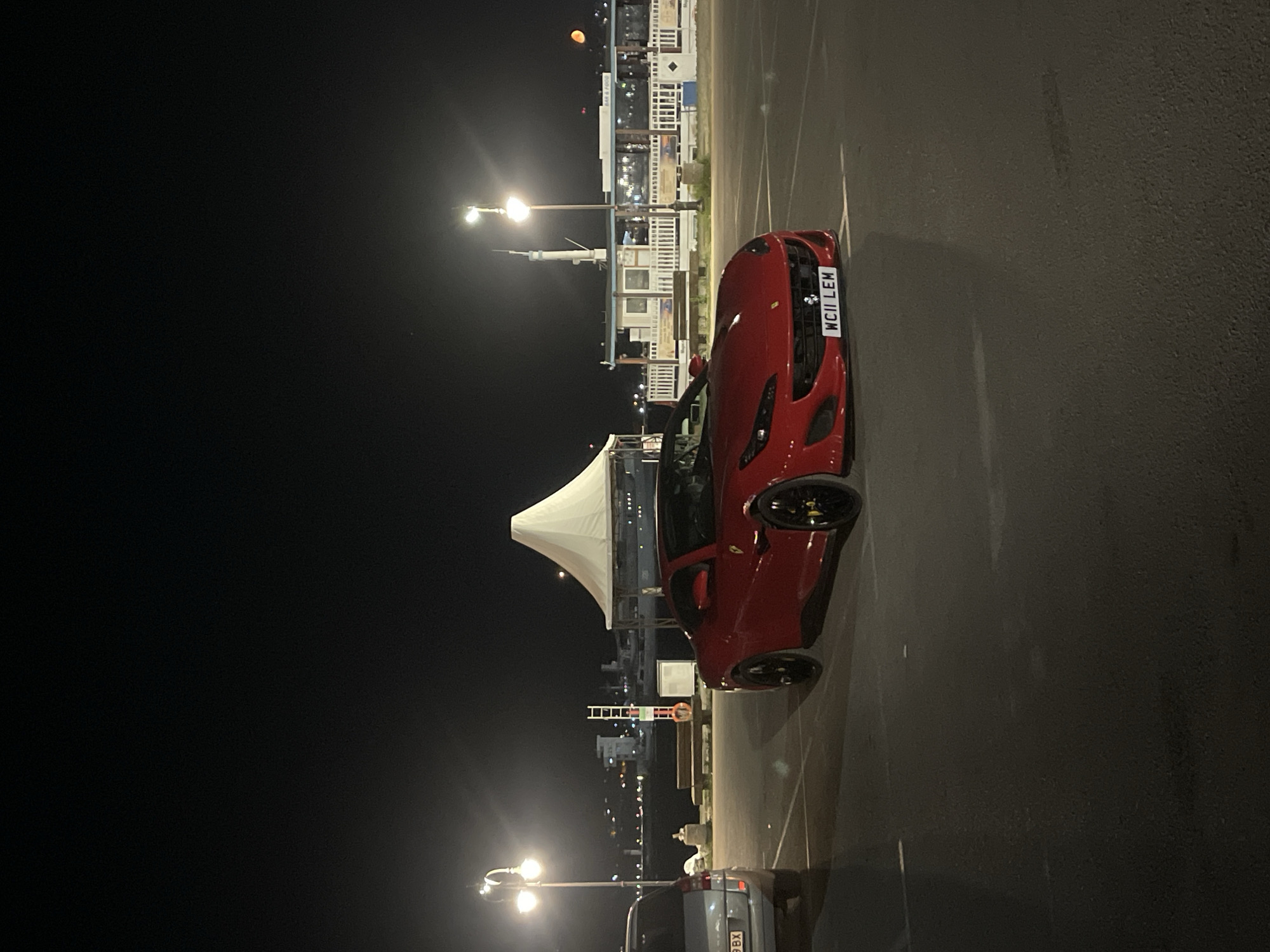 A red Ferrari Portofino is parked in a lot at night. The car is facing forward, with its headlights on. The license plate reads "WCII LEM". In the background, there is a waterfront area with buildings and lights. A white tent is set up to the left of the car, and a silver SUV is parked to its left. The location appears to be near the Varna Sea Garden, a prominent park and cultural center in Varna, Bulgaria. The scene is illuminated by streetlights and ambient light from the surrounding area. There are no people visible in the image.