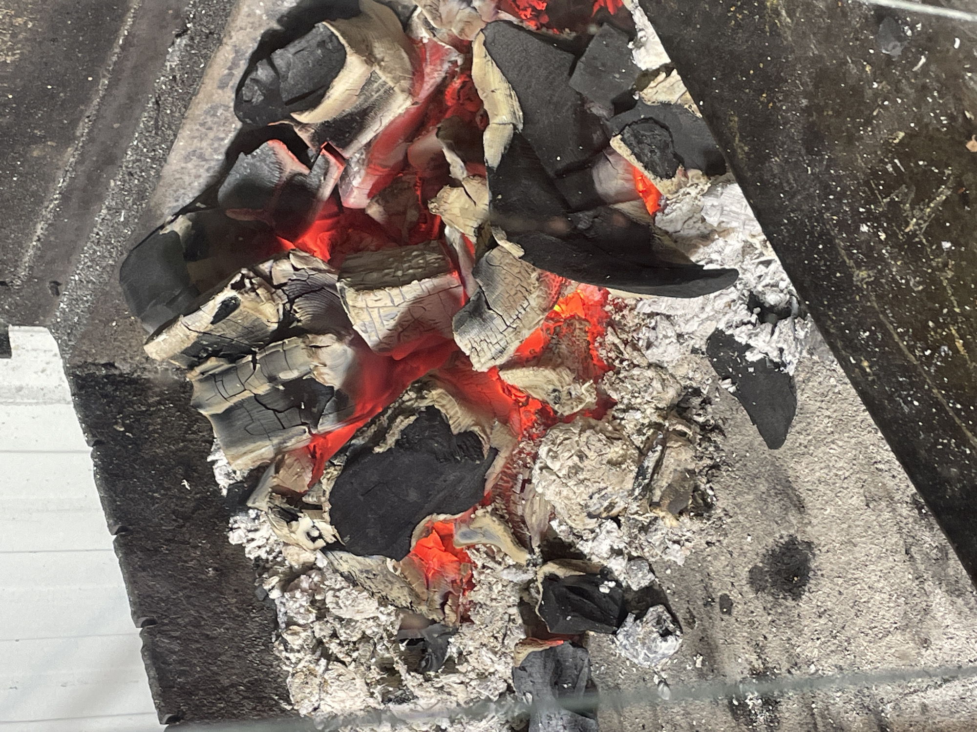 The image shows a close-up, high-angle view of hot coals and ash in a grill. The coals are primarily black, with some glowing red, indicating they are still hot. Various pieces of wood charcoal, some partially burned and covered in white ash, are piled together. The grill appears to be made of dark metal and is situated outdoors, possibly in a backyard or patio area. The background shows a textured, light-colored surface, likely a wall or fence. There are no people or animals visible in the image.