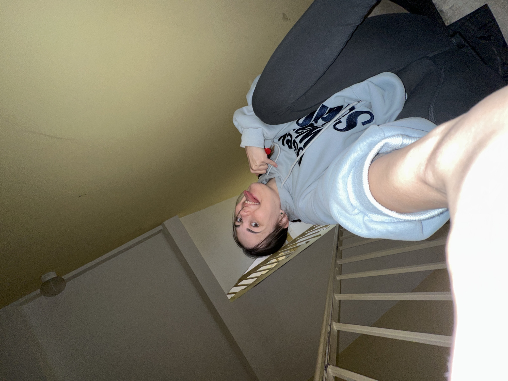 A young woman is pictured in a low-angle selfie. She is sticking her tongue out and holding a red object. She is wearing a light blue hoodie with dark letters and dark grey leggings. The setting appears to be inside a stairwell. The walls are light yellow and the stairs are made of light-colored wood. This photo was taken in Perm, Russia.