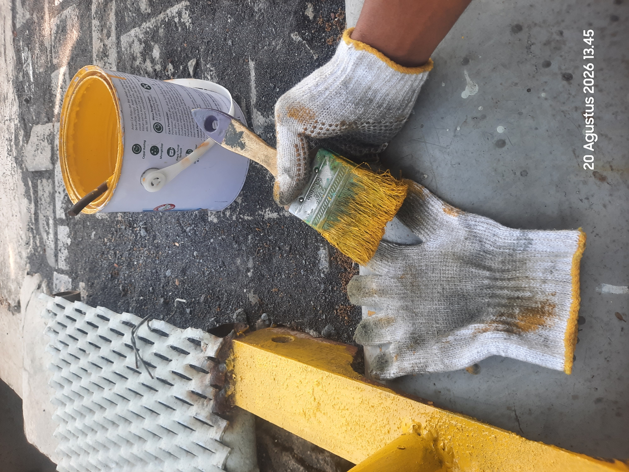 In Gending, Indonesia, a close-up shot shows a person's hands, protected by white and grey work gloves with yellow trim, preparing to paint. One hand is holding a paintbrush with yellow bristles, which appears to have yellow paint on it. The paintbrush is positioned over a can of yellow paint, which also contains a stirring stick. The can has text and diagrams on its label. In the background, a yellow metal beam is visible, with rust and drips of yellow paint on it. To the left of the beam is a white, grated metal structure. The ground is a mixture of asphalt, gravel, and concrete, with some scattered white paint splatters. A timestamp in the bottom right corner reads "20 Agustus 2026 13.45".