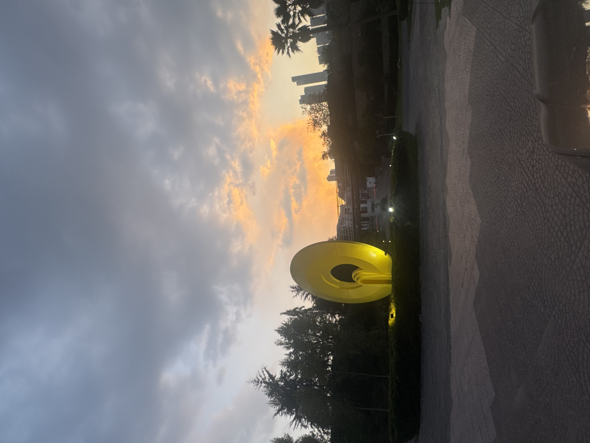 The image captures a late-day scene in Huixquilucan de Degollado, Mexico. In the foreground, a large, circular, yellow public art sculpture stands illuminated against a backdrop of green trees. The sky is divided into grey clouds on the left and a sunset gradient of orange and yellow on the right. In the mid-ground, buildings and skyscrapers can be seen in the distance, with palm trees punctuating the skyline. A paved walkway or road extends into the distance on the right side of the frame. No people are immediately visible.