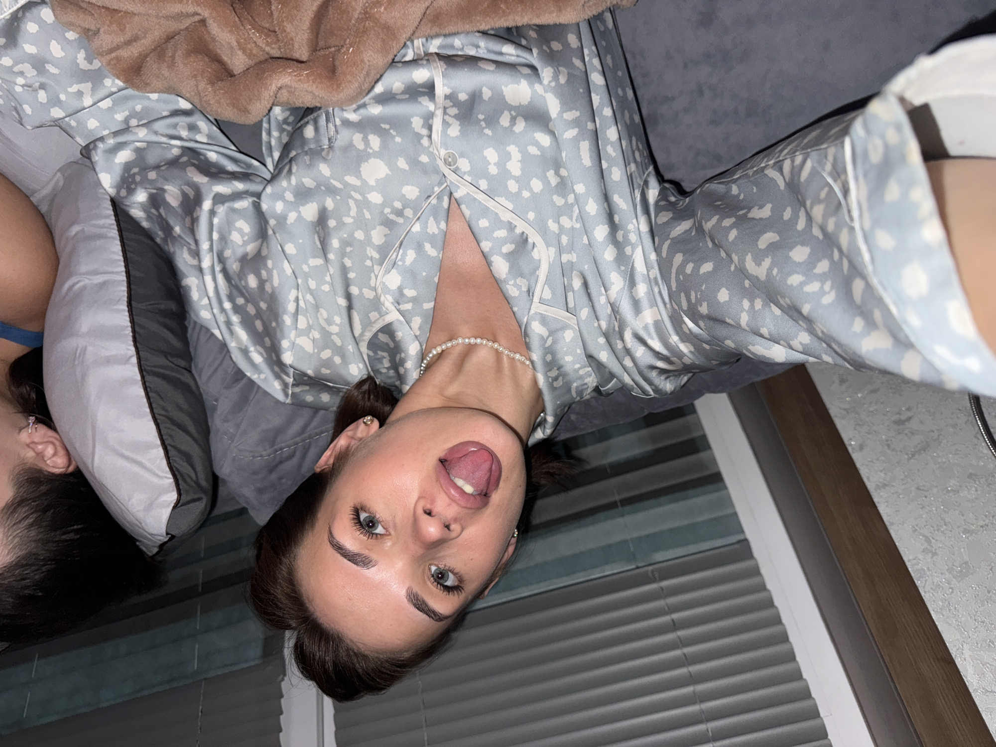 The image shows a woman with dark hair, light skin, and blue eyes lying on a bed. She is wearing a blue pajama top with a white pattern and a pearl necklace. She is sticking her tongue out and looking at the camera. Behind her is a second person partially visible. They are lying on a pillow. The background includes window blinds and a patterned wall. The image appears to have been taken indoors. It is not possible to confirm that the image was taken in the city of Lobanovo, Russia from this image alone.