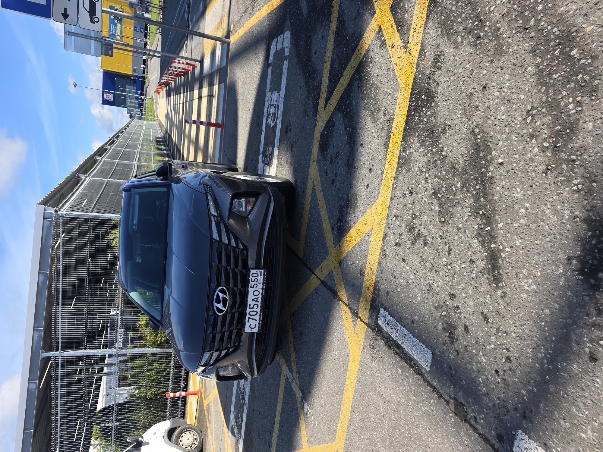 A dark gray Hyundai Tucson SUV is parked in a parking lot on a sunny day. The car is facing forward and is slightly angled to the left. The SUV has a license plate that reads "C705AO 550". The ground is asphalt and is marked with yellow lines. There is a white pedestrian crossing in front of the car, and a white symbol of a person in a wheelchair is painted on the ground to the right of the car. In the background, there is a metal fence and a building with a blue and yellow facade. There are also traffic cones in the background. The sky is blue with some clouds.