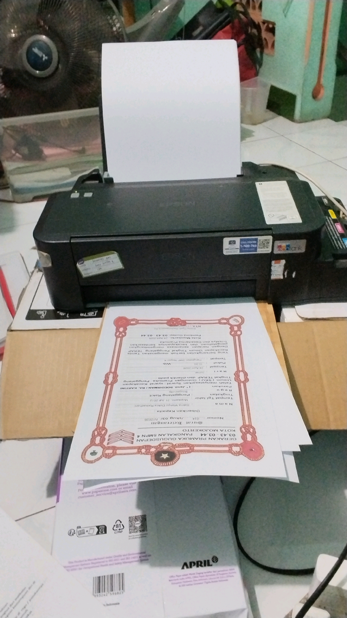 A high-angle, medium shot captures a printer on a tiled floor. The printer, black and rectangular, sits centrally, with a white sheet of paper protruding from its top. The paper displays text and a decorative border. To the left of the printer, a large, black electric fan stands, its grill partially visible. In the background, out of focus, a pale green wall is present, with various objects including what appears to be a light blue cooler, wooden spoons, and electrical equipment. The scene is lit by ambient light, with no direct illumination source evident. The floor tiles are light-colored and rectangular. The overall impression is of a home or office environment.