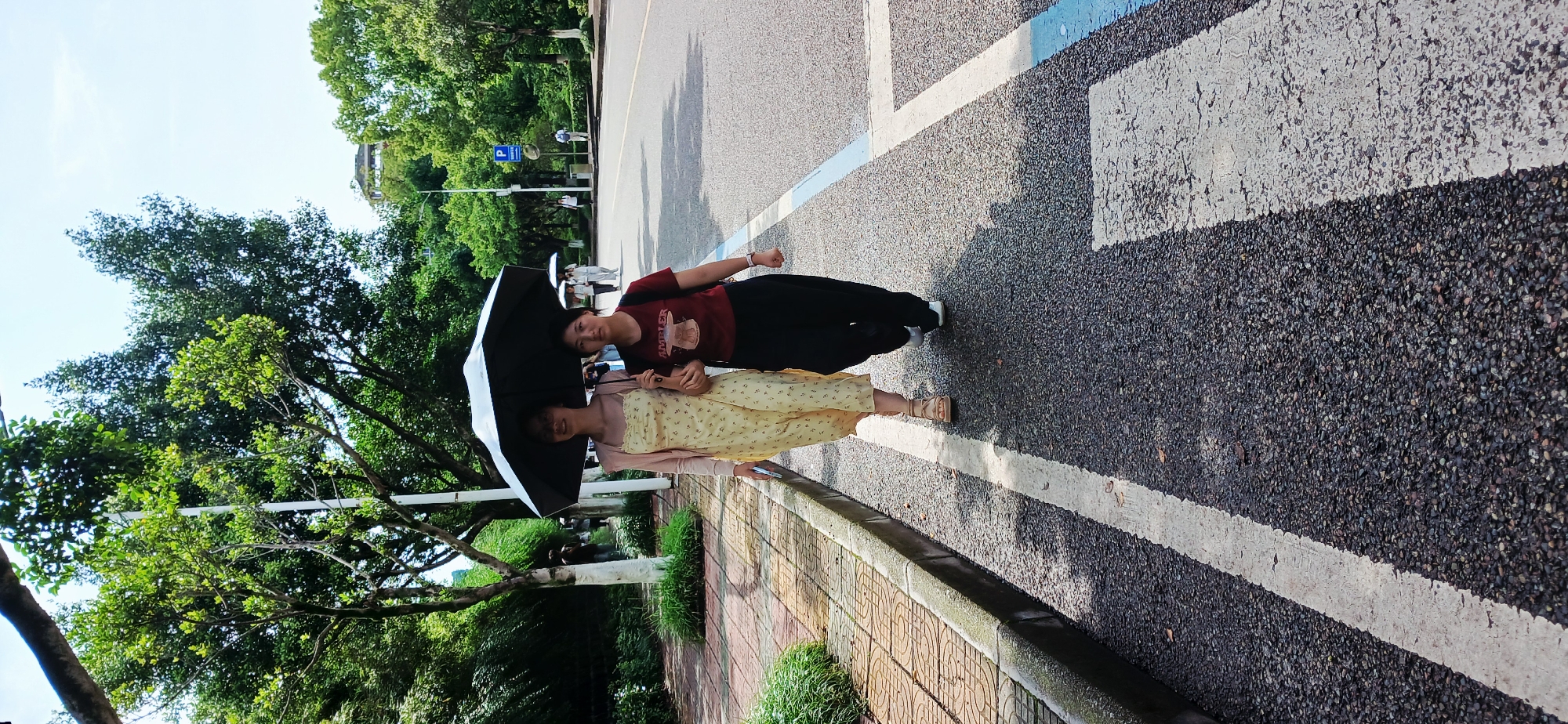 Two females are standing under an open umbrella on an asphalt surface in Chongqing High-tech Zone, China. One female wears a yellow dress and sandals; the other wears a maroon shirt and black pants. The ground is marked with white and blue painted stripes. Trees are visible in the background. A concrete curb and a strip of vegetation border the edge of the asphalt.