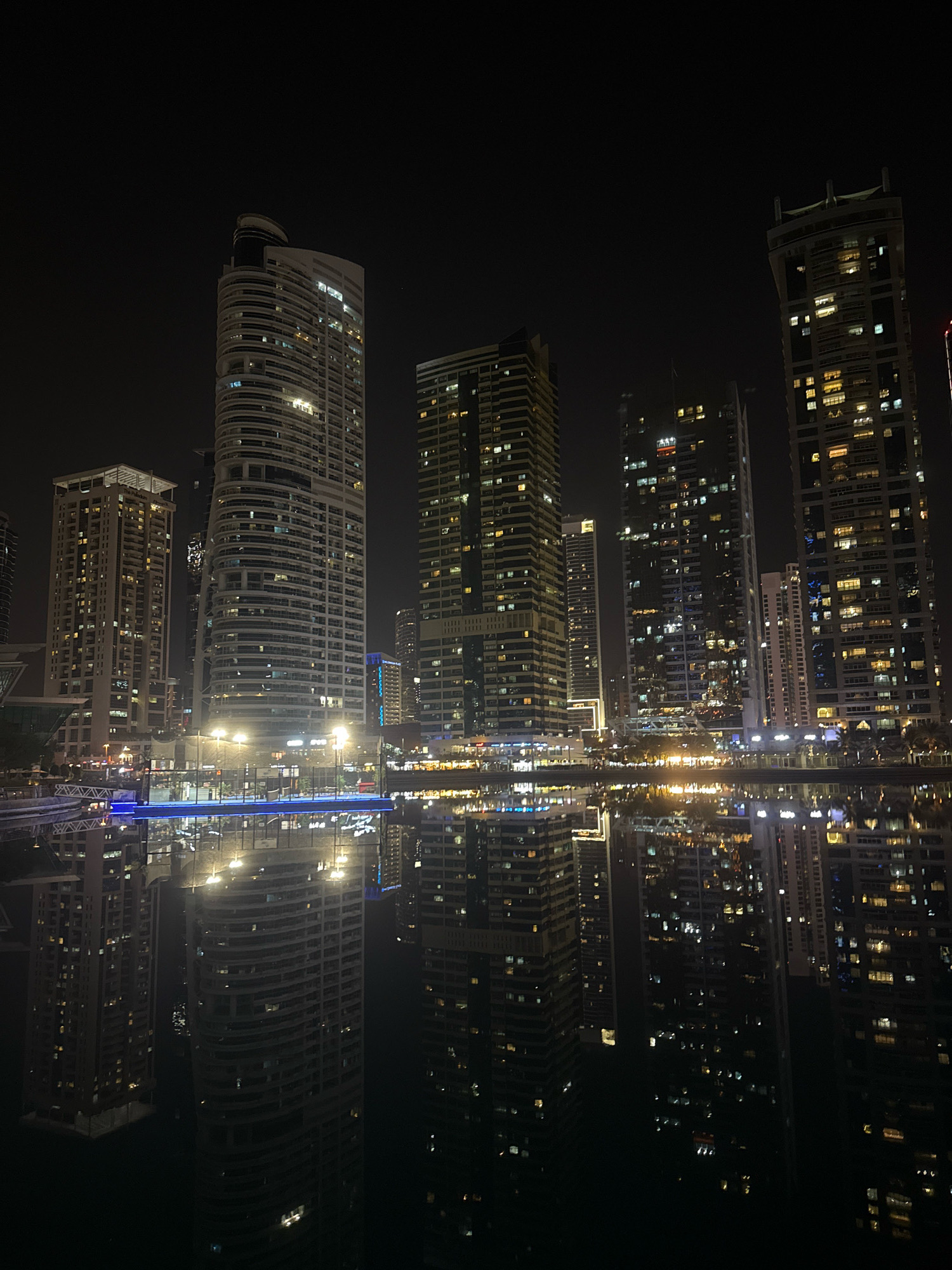 This is a nighttime photograph of the Dubai Marina, in Dubai, United Arab Emirates. The image captures the illuminated skyscrapers reflecting on the still water of the marina. The foreground features the dark water with a clear reflection of the buildings and lights above. The middle ground shows a brightly lit walkway or platform over the water, with several lampposts providing illumination. In the background, numerous high-rise buildings of varying architectural styles, all lit with windows, dominate the skyline. The sky is dark and clear. There are no people or vehicles visible in the frame. The overall impression is one of a vibrant, modern urban landscape at night.