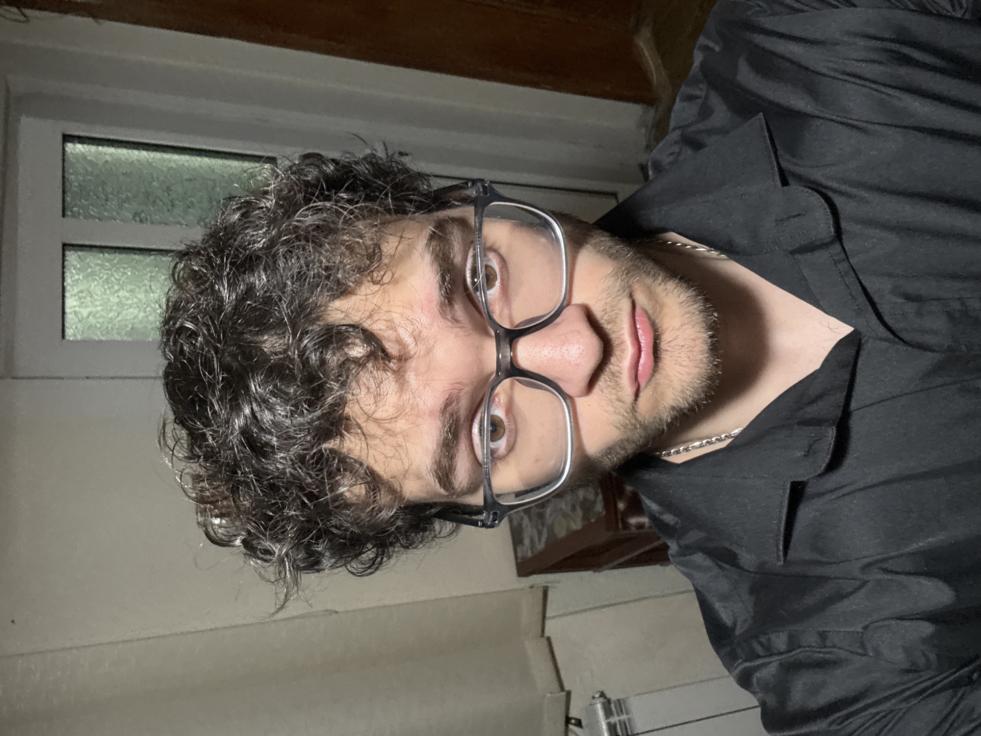 A medium-close-up shot shows a man with curly dark hair and stubble wearing clear-framed glasses and a black button-down shirt. He is looking directly at the camera. A silver chain is visible around his neck. Behind him, there is a window with frosted glass panes and a dark wooden ceiling. The lighting appears to be artificial, casting shadows on the wall. The scene lacks elements to establish a specific location in Ilindentsi, Bulgaria.
