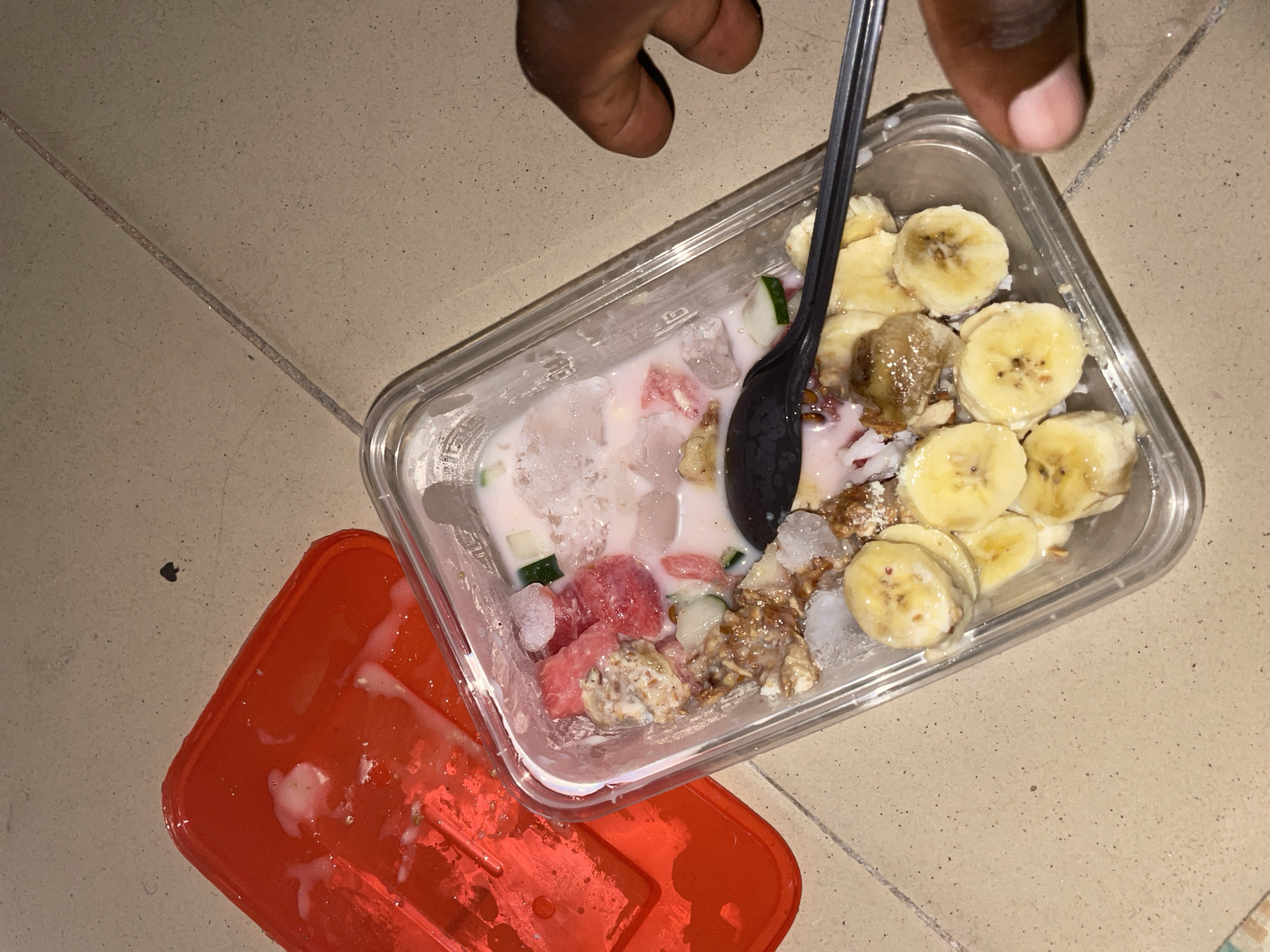 A close-up, high-angle overhead shot depicts a clear rectangular plastic food container resting on a light-colored tiled surface in Jalingo, Nigeria. The container holds a dessert or snack mixture featuring a milky, light pink liquid base with visible chunks of ice. Ingredients within the container include numerous slices of yellow banana arranged along the bottom edge, irregular pieces of red fruit (consistent with watermelon) and diced green vegetable (consistent with cucumber) suspended in the liquid, and a brown, granular, lumpy mixture (possibly granola or crushed nuts) towards the upper left portion. A black plastic spoon is positioned diagonally within the container, with its bowl submerged in the mixture and its handle extending towards the right. To the left of the container, a red plastic lid, slightly larger than the container, rests upside down with milky residue visible on its interior surface. A dark-skinned human hand is partially visible on the right side of the frame, with fingers gently curled around the lower right exterior of the clear container. The background consists of light brown or beige floor tiles with visible grout lines and scattered dark specks. A distinct small black spade-shaped mark is visible on the tile surface in the upper left quadrant of the frame.