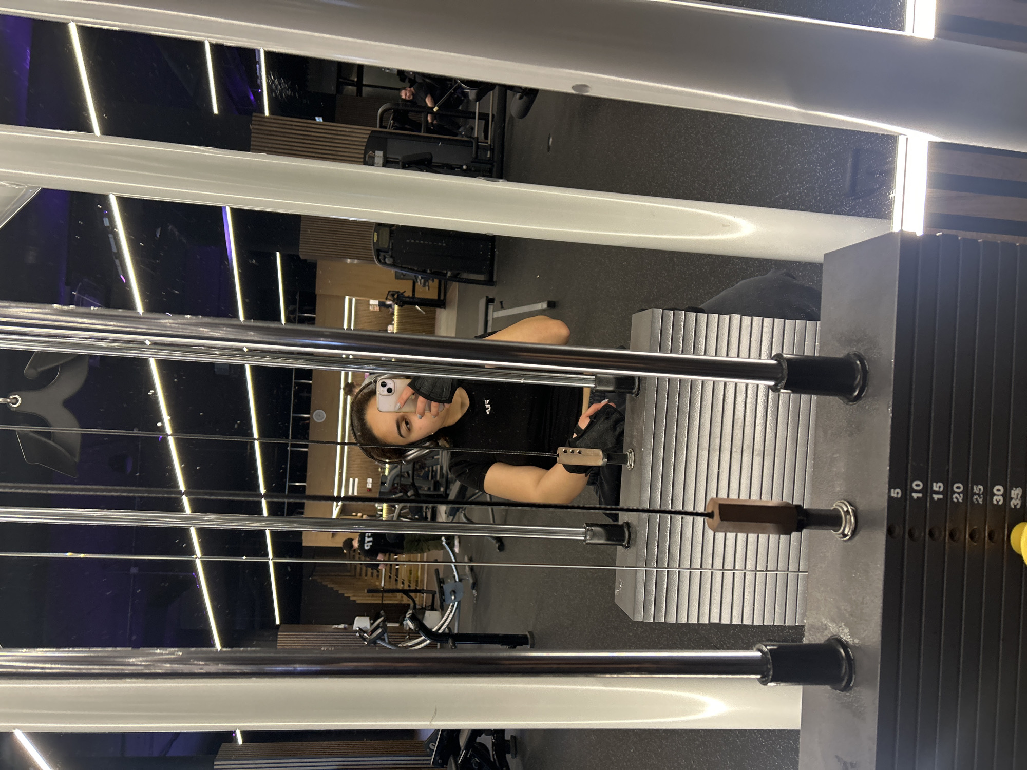 A young woman is captured in a mirror selfie within a modern gym in Burgas, Bulgaria. She is seen from the chest up, holding a white smartphone to take the picture. She wears a black short-sleeved athletic top with a white "AUF" logo, black fingerless gloves, and black athletic pants. Her dark hair is pulled back, and she has visible makeup. She appears to be standing or sitting beside a cable exercise machine.
In the foreground, various polished metal cables and bars of the gym equipment are visible, creating horizontal and vertical lines across the frame. To the right, a black weight stack for the machine displays numerical increments: 5, 10, 15, 20, 25, 30, 35, with a yellow pin inserted, indicating a selected weight. A wooden handle is attached to one of the cables near her right hand.
The reflection in the mirror reveals a spacious, contemporary gym setting. The background features dark walls, some with a subtle texture, and sections with horizontal wooden slats. The gym is well-lit by modern linear LED lights, with some emitting a bright white glow and others a soft purple or blue hue, contributing to a sleek aesthetic. Other gym equipment, including what appears to be a treadmill and benches, is reflected in the distance. Another person is visible in the background reflection, seated on a piece of equipment, wearing a dark shirt with partially visible text, possibly "TLB". The overall atmosphere is clean, well-lit, and focused on fitness activities.
