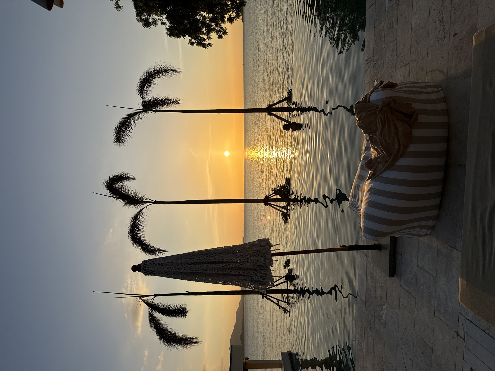 The image depicts a tranquil sunset scene at a resort in Koh Samui District, Thailand. The foreground features a paved poolside area with a large, striped beanbag chair covered by a tan towel. Adjacent to the beanbag, a partially open, crochet-style umbrella stands prominently. Beyond the pool's edge, the calm ocean stretches towards the horizon, reflecting the warm hues of the setting sun. Three silhouetted palm trees stand in the water, their trunks anchored to the seabed, adding to the tropical ambiance. The sun, a bright orb, is setting over the ocean, casting a golden glow and a shimmering reflection on the water's surface. In the distance, a hazy coastline is visible. The sky transitions from a pale blue at the top to a warm yellow and orange gradient near the horizon. The overall atmosphere is serene and idyllic.