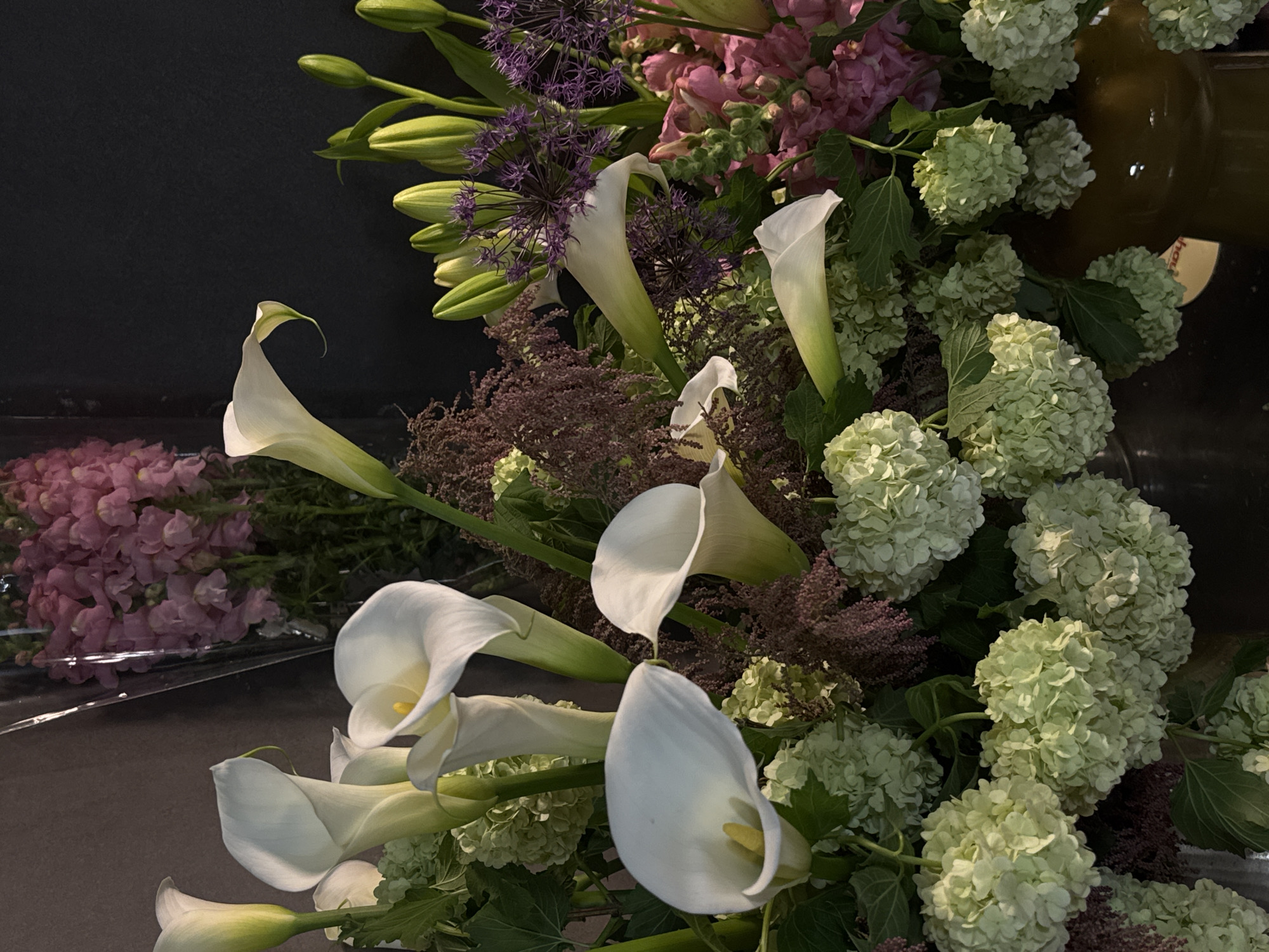 This vertical close-up image showcases a vibrant and varied floral arrangement, likely set in a florist shop or studio in Yerevan, Armenia. The composition is dominated by an abundant display of fresh cut flowers against a dark, featureless background, which accentuates their colors and forms.
The primary subjects are numerous white calla lilies (Zantedeschia aethiopica), characterized by their elegant, trumpet-shaped spathes and yellow spadixes, prominent throughout the foreground and mid-ground. Interspersed among them are clusters of light green, rounded florets, likely hydrangeas or snowball viburnums, adding a lush, textured element. Deep purple, spiky spherical flowers, resembling alliums or ornamental onions, provide a striking color contrast and unique texture. Complementing these are feathery bunches of brownish-purple filler flowers, possibly astilbe. In the background, bunches of soft pink or mauve clustered flowers, possibly snapdragons or stock, are visible, some still wrapped in clear plastic, suggesting they are newly arrived or awaiting arrangement.
There is no specific activity depicted; the image is a still life, focusing on the beauty and variety of the floral selection. The lighting appears artificial and directed, highlighting the petals' textures and creating subtle shadows that give depth to the arrangement. A dark brown or bronze vase is partially visible on the right side of the frame, blending into the dark background. A small yellow tag with the partial text "ho" is visible near the vase. The overall impression is one of elegance and natural beauty, with a rich mix of shapes, colors, and textures.