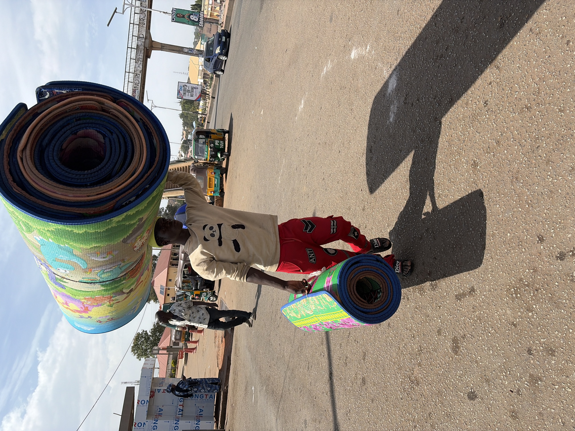 A bustling street scene, likely in Jos, Nigeria, is captured on a bright, sunny day with a clear sky and scattered clouds. The image's primary subject is a man carrying a large, heavy bundle of colorful, patterned rolled mats over his left shoulder and a smaller stack in his right hand. He wears a light-colored hooded sweatshirt with a panda graphic, red pants with the word "ANTI" on them, and "Dior" branded sandals.
The man appears to be a vendor or transporting goods, possibly walking along the asphalt road. Strong shadows indicate either mid-morning or afternoon. In the background, other people are visible, along with several vehicles including green and yellow tuk-tuks (locally known as keke napep) and a blue car. One tuk-tuk has "PTA 7224" visible on its rear.
An overhead pedestrian bridge crosses the road, with the inscription "INDIRISVADBRISCE" along its structure. Various signs and billboards line the street, featuring prominent text such as the political slogan "TINUBU -OR- NOTHING," the logo for "Opay" (a mobile money service), and a banner with "Shields CTRER CAF" (likely Cyber Cafe). At the bottom left, bundles of corrugated metal sheets, likely building materials, are stacked, bearing partial texts like "RON UILDIN," "RONG," "ONGTA," and "GTALRONG." The scene conveys the vibrant activity of a Nigerian urban area.