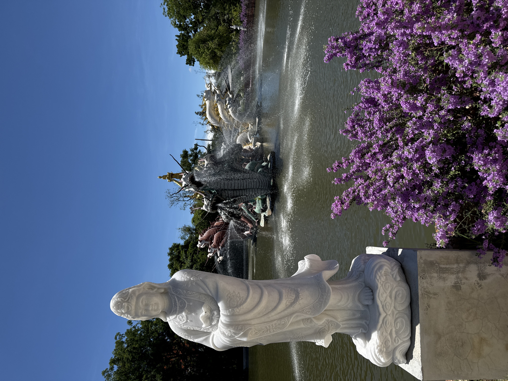 This image depicts a statue of a woman in flowing robes. The statue is white and appears to be made of marble or a similar material. It stands on a decorative base. Behind the statue is a pond or lake with a fountain. The fountain consists of multiple sculpted dragons and other figures. Water is spraying from the fountain. Purple flowers are visible to the right of the fountain. The sky is clear and blue. This is likely a tourist attraction in Bang Pu Subdistrict Municipality, Thailand.