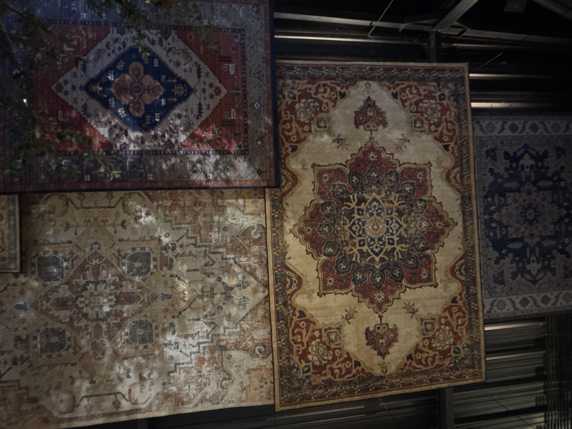 The image shows a collection of hanging rugs in the Ubud District of Indonesia. Four rugs are visible, each with distinct patterns and colors. The top left rug features a red and blue geometric design. Below it, another rug displays a lighter-colored, more intricate pattern with elements of blue and red. To the right of these, a beige rug with a complex red and blue floral or mandala-like design hangs. Further to the right is a rug with predominantly blue patterns. The rugs appear to be displayed in an interior space, possibly a shop or market, with metal framework visible in the background.