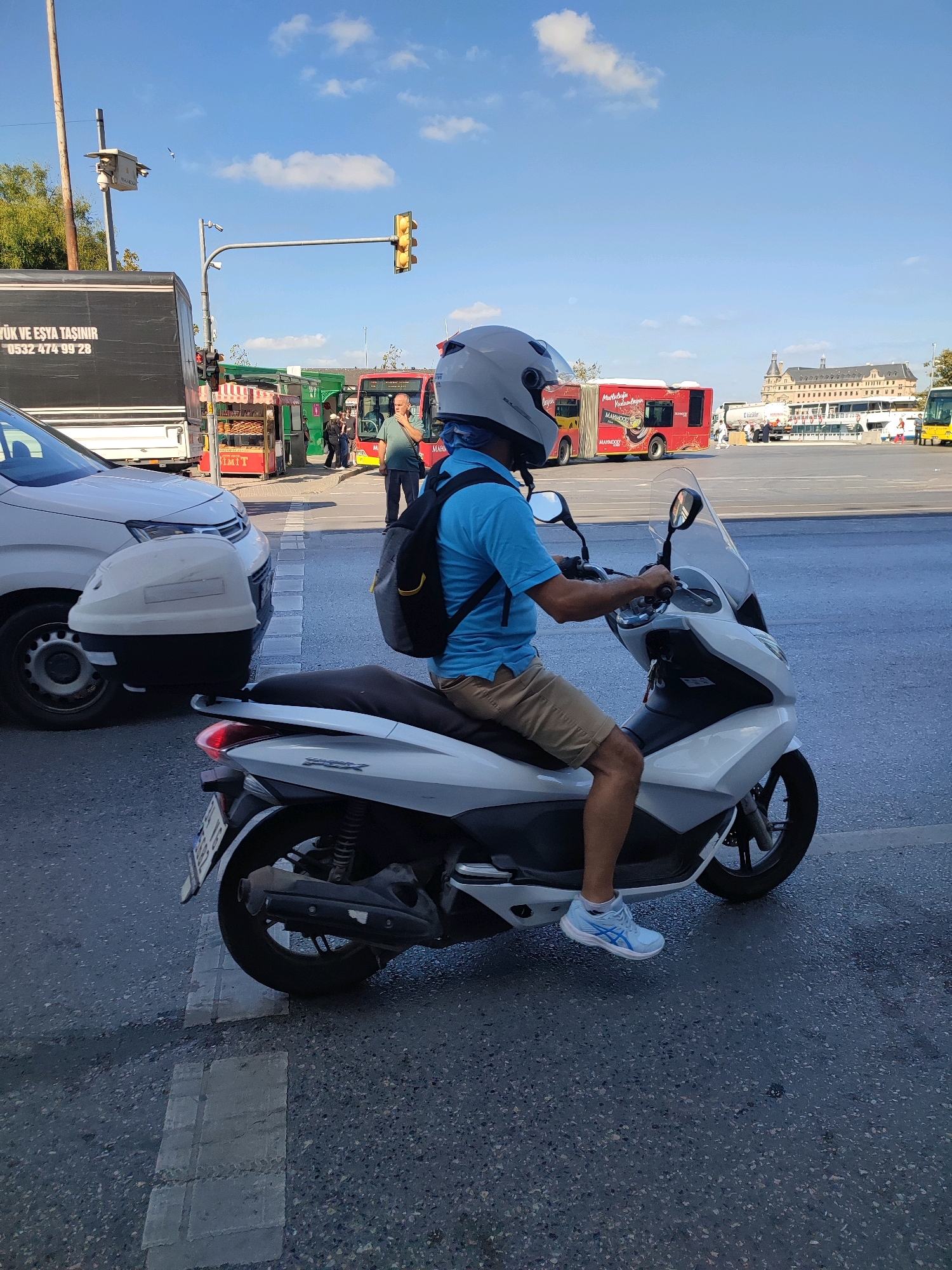 A man wearing a blue shirt, beige shorts, and a white helmet is riding a white scooter on a city street. He has a black backpack on his back. The scooter has a clear windshield and a license plate. In the background, there is a white car, a red bus, and a large building. A traffic light is visible in the upper part of the image. The sky is blue with some clouds. The scene appears to be in Kadıköy, Turkey, likely near the Haydarpaşa Train Station due to the distinctive building in the background. The street is paved and appears to be a main thoroughfare with multiple lanes.