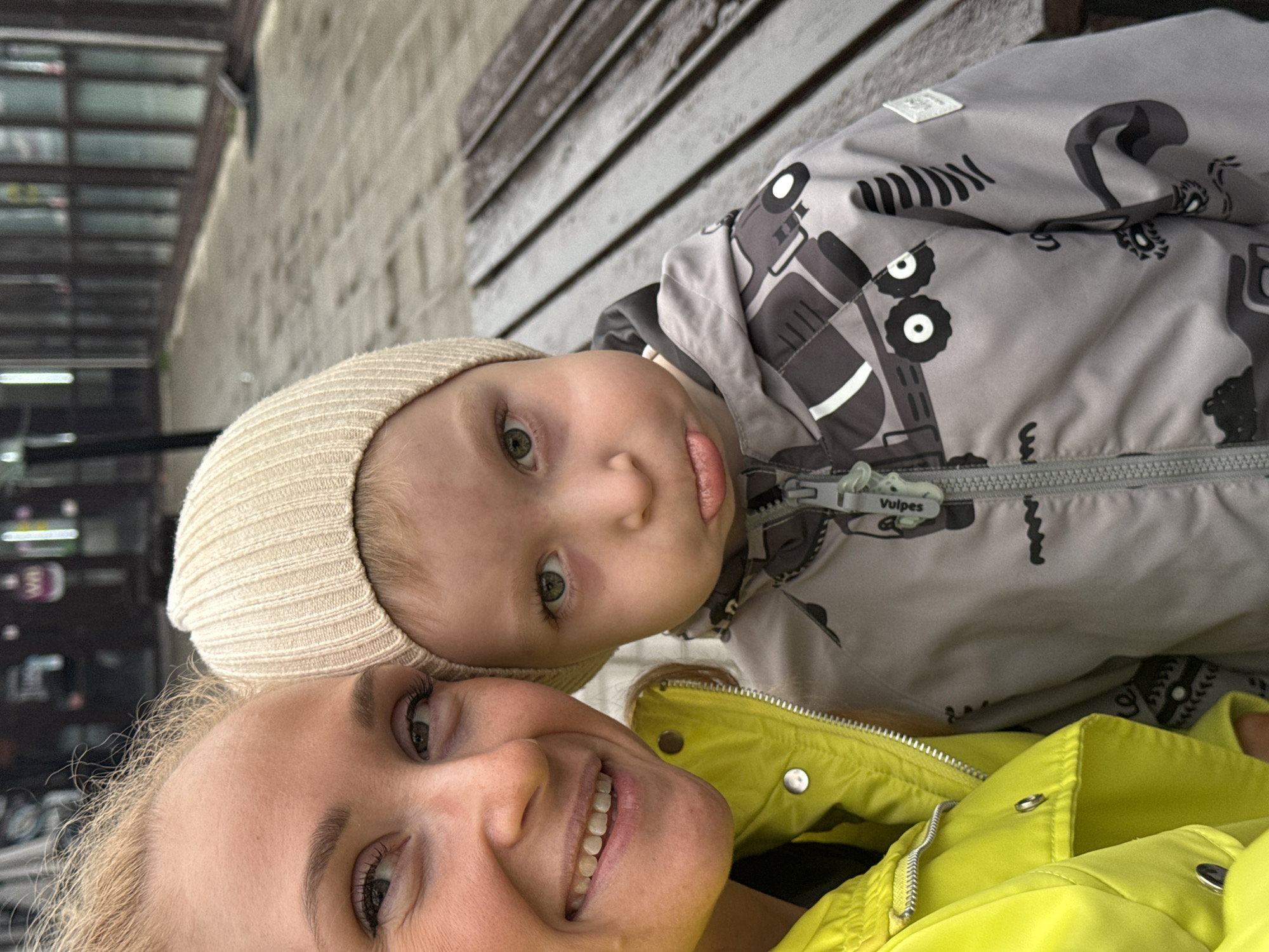 A portrait-oriented, close-up selfie features a smiling woman and a young child, appearing to be outdoors or in a sheltered urban area.
The woman, in the lower left of the frame (when upright), has blonde hair and wears a bright yellow zip-up jacket with silver snaps. Her right eye, nose, and mouth are visible as she smiles towards the camera, conveying a happy expression.
Above and to her right is a young child, likely a toddler, wearing a light beige knit beanie and a grey jacket patterned with black and white stylized vehicles, including what look like tractors and cars. The child has green/hazel eyes and looks directly at the camera, with their tongue slightly extended, conveying a playful or curious expression. On the child's jacket zipper pull, the text "Vulpes" is clearly visible.
The background is blurred, with a wooden slatted surface visible behind the child and an indistinct, darker area with shelves or structures behind the woman, suggesting an urban environment, possibly a covered walkway or near a building. The lighting indicates it is daytime, possibly overcast or within a covered space due to the soft light and absence of harsh shadows.