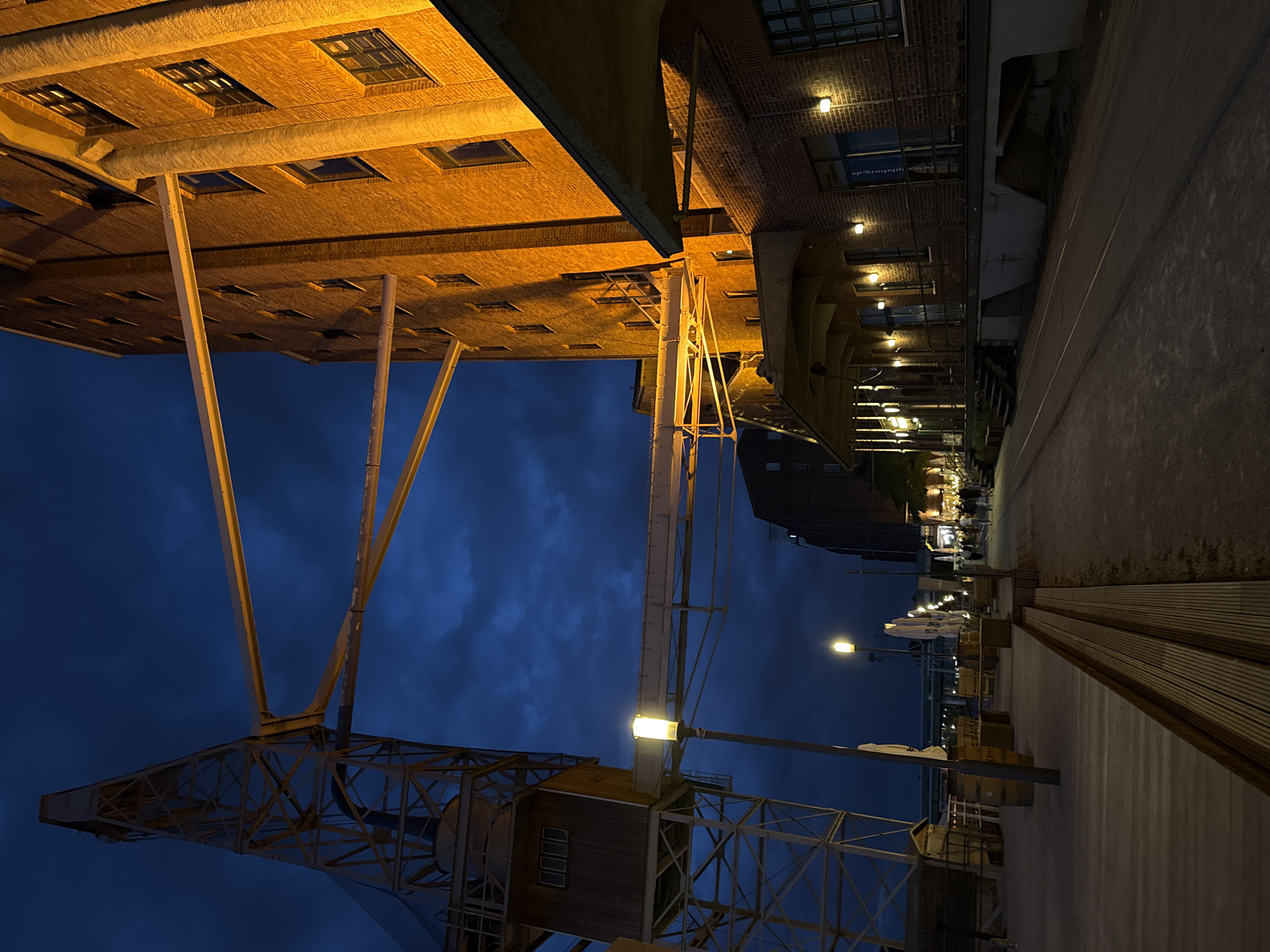 This image captures an evening scene in the city of Duisburg, Germany, likely at the Marina Landschaftspark. On the left, a towering yellow industrial crane dominates the frame, its intricate metal structure silhouetted against a dark blue, cloudy sky. A wooden cabin is visible partway up the crane. Extending horizontally from the crane are yellow metal beams, some of which connect to a large, multi-story brick building on the right. This building is illuminated by warm yellow lights, highlighting its brick facade and numerous windows.
In the foreground, a wooden walkway with built-in benches lines the scene. A streetlamp with a glowing yellow light stands on the walkway. Further down the walkway, several white market umbrellas are visible, suggesting an outdoor seating area or market. Beyond the umbrellas, a street or path runs parallel to the brick building, where a few people can be seen walking. The ground appears to be a mixture of gravel and what looks like railway tracks. The overall atmosphere is one of twilight industrial heritage, with the imposing structures illuminated against the darkening sky.