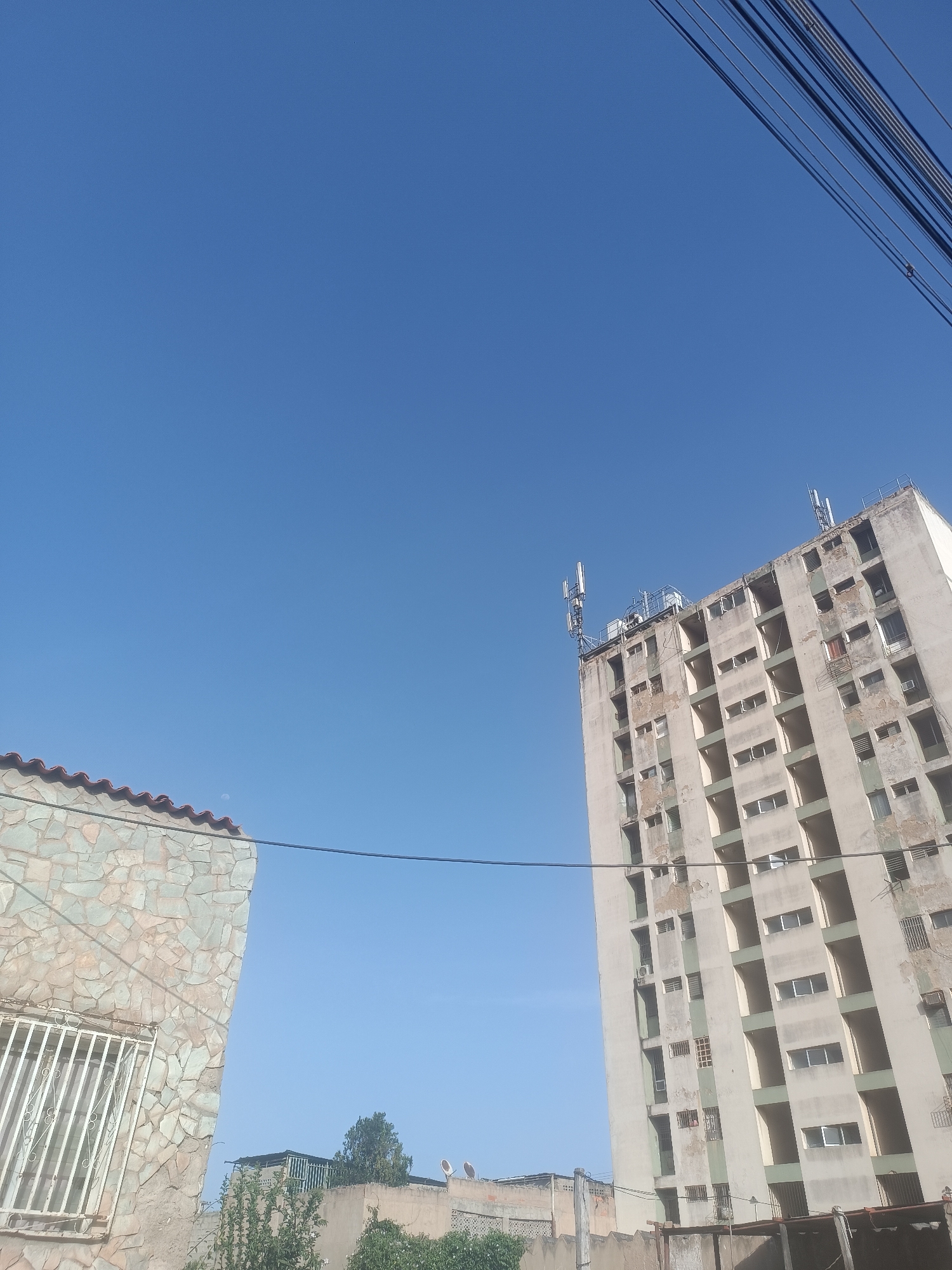 The image depicts a view of a city scene, likely in Porlamar, Venezuela, based on architectural styles. The sky is clear and blue. Electrical wires traverse the upper right corner of the frame.
To the left is a portion of a single-story building with a tiled roof. The walls are clad in stone. A barred window is visible.
To the right, a tall, weathered, multi-story building dominates. It appears to be residential, with multiple windows and balconies. The building shows signs of age and disrepair, with faded paint and visible wear. Various antennas are mounted on its roof. The top of this building is located in the top right of the frame.
In the foreground, partial views of other buildings and some vegetation are visible. A fence is also present.
