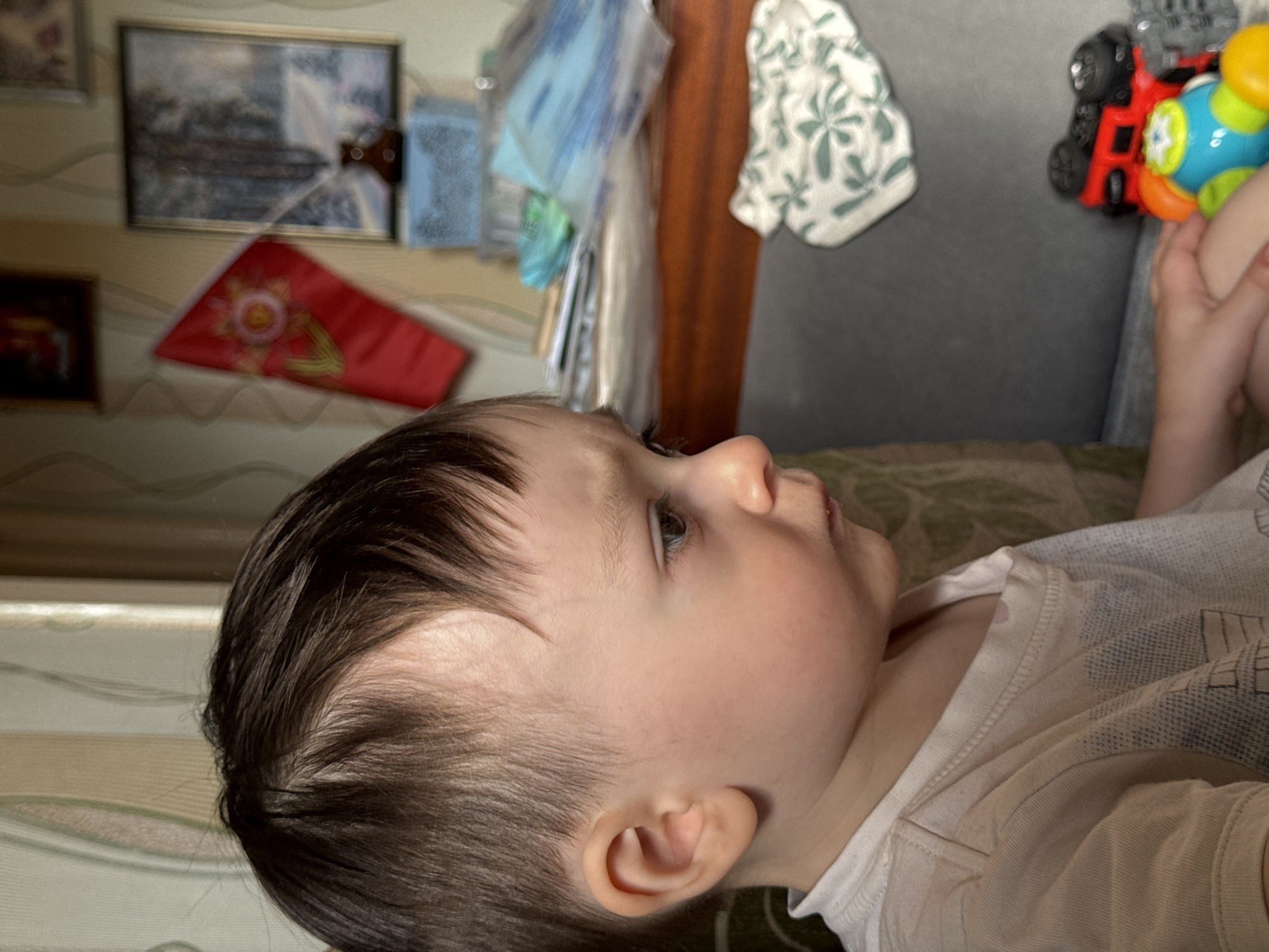 A close-up, tilted, indoor shot features a young child lying on a surface covered with a patterned fabric. The child has dark brown hair and fair skin, and is facing right, with their head resting on the surface. The child is wearing a white t-shirt and appears to be holding a colorful toy with red, gray, blue, and yellow elements in their hands.
In the background, a wall is visible, adorned with several framed items, including a landscape painting and a red banner. To the right, behind the child, is a gray surface with a white patterned cloth hanging from it. Behind the wall appear to be hanging bags or clothes, and there are wooden interior components. The photo does not reveal any information about a location in the city of Izhevsk, Russia.