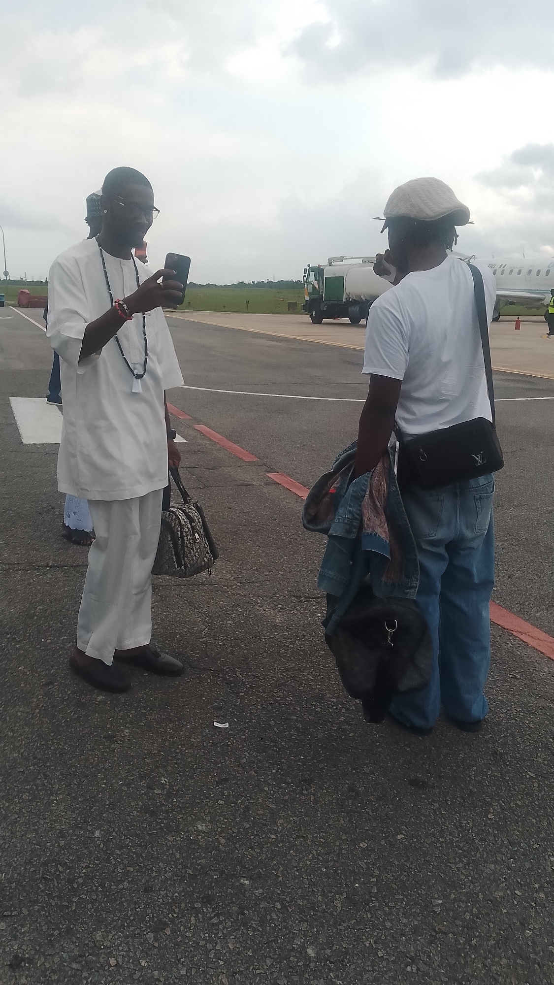 Two individuals are on an airport tarmac in Orerokpe, Nigeria, under an overcast sky. The person on the left is a man wearing a white tunic and pants, with a beaded necklace and a bracelet. He is holding a smartphone and appears to be taking a picture or video. He is also carrying a designer handbag. The person on the right, facing away from the camera, is wearing a white t-shirt, blue jeans, and a hat. They are carrying a denim jacket over their arm and a black crossbody bag. In the background, an airplane and a fuel truck are visible. The tarmac is asphalt with red markings.