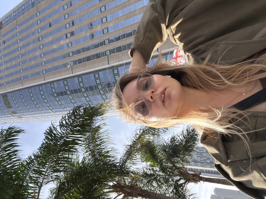 An image taken from a low angle captures a female individual posing in an urban environment. The subject, a woman with long, windswept blonde hair, is centered in the lower half of the frame. She wears clear-framed glasses with subtly tinted lenses, a white earbud in her right ear, and a golden necklace with a small pendant. Her mouth is slightly open, and she looks directly at the camera. She is dressed in an olive-green zipped jacket over a darker top. Her right hand is raised, partially obscured by her hair on the right side of her head.
In the background, on the left, several palm trees with green fronds are visible against a clear blue sky. Dominating the upper and right portions of the background is a modern, tall skyscraper featuring a curved facade of blue-tinted windows and light-colored vertical panels. To the left of this main skyscraper, another less distinct, tall building can be seen. Below the main skyscraper and behind the woman's right shoulder, a flag is partially visible, identifiable as the national flag of Georgia, depicting a large red cross on a white background with four smaller red crosses in the quadrants.
The scene is illuminated by natural light, indicative of a sunny day. The location is in Tel-Aviv, Israel, specifically in the vicinity of the Azrieli Sarona Tower, near the Embassy of Georgia at Leonardo Da Vinci Street 21.