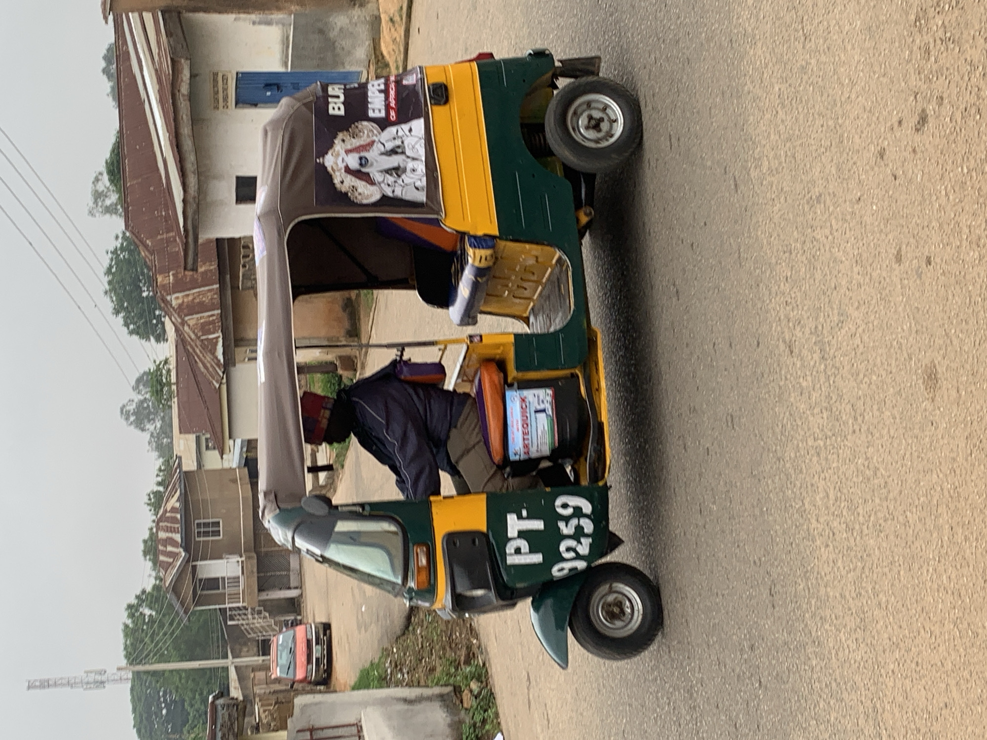 The image depicts a yellow and green auto-rickshaw, commonly known as an "okada" or "Keke Napep," driving on a paved road in Jos, Nigeria. The auto-rickshaw is adorned with a large poster featuring a regal figure, identified as "BUR EMPIRE OF AFRICA." The driver, wearing a dark jacket and a red and multi-colored hat, is visible behind the steering wheel. The background shows residential buildings with corrugated iron roofs, typical of Jos architecture, and a telecommunications tower in the distance. The scene suggests a typical urban streetscape in Jos.