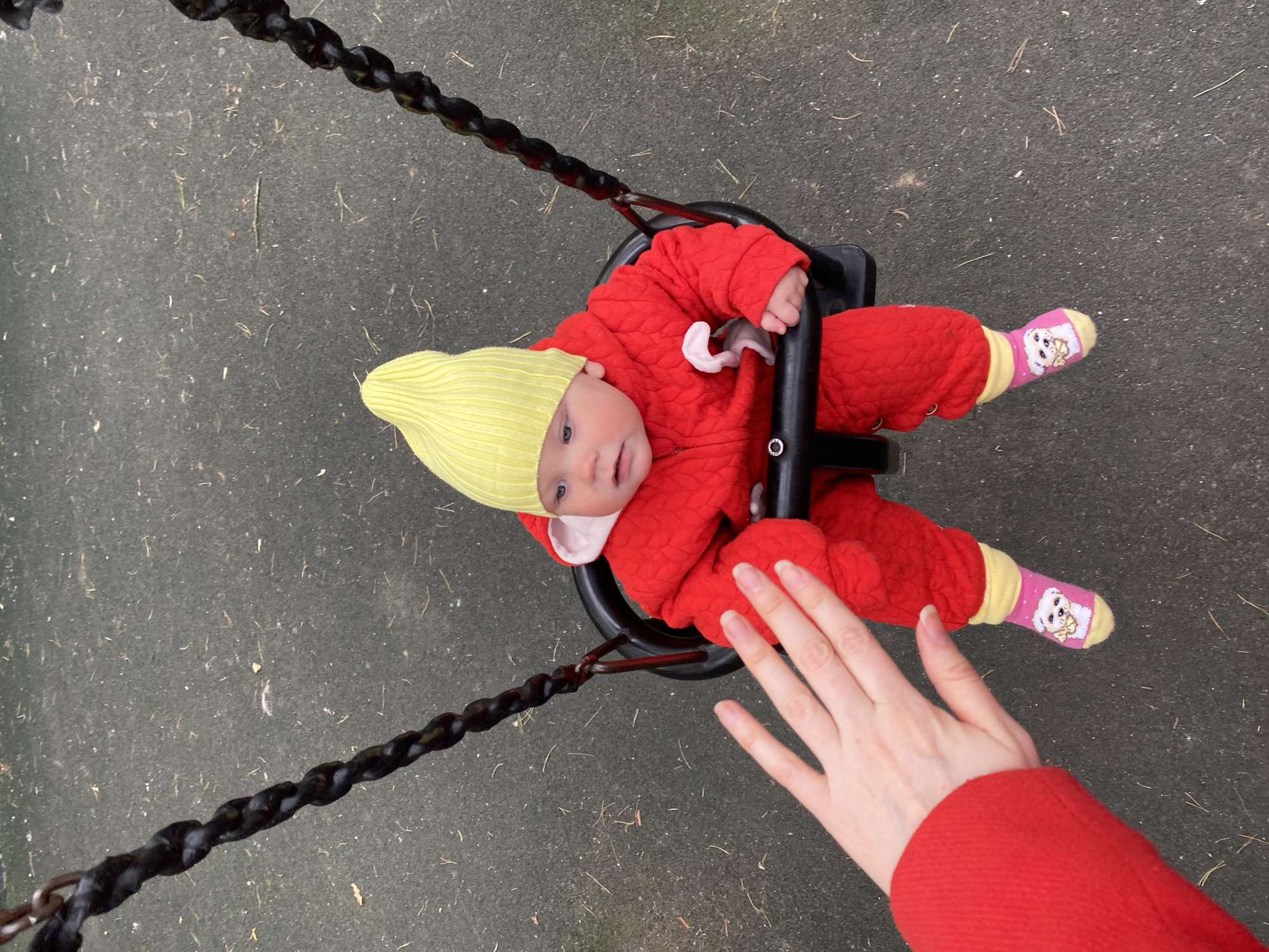 An overhead, high-angle shot captures a baby in a swing at an outdoor playground, likely in Yekaterinburg, Russia. The infant is dressed in a vibrant red quilted snowsuit and a bright yellow knitted beanie, looking upwards with wide, curious eyes and a slightly open mouth. Pink and yellow socks, featuring an image of a white dog, are visible on the baby's feet.
The baby sits in a black plastic swing seat, suspended by chains covered in black protective sheathing. An adult's fair-skinned hand, clad in a red sleeve that appears to match the baby's outerwear, is positioned in the foreground near the baby's feet, suggesting interaction, possibly during the act of pushing the swing or adjusting the baby.
The ground beneath the swing is a dark gray or black rubberized safety surface, typical of playgrounds, with scattered small bits of debris such as dried leaves or twigs. The lighting is soft and diffused, indicating an overcast day with no harsh shadows. The warm clothing of the baby suggests cooler weather. The image conveys a playful and caring moment. There is no visible text in the media file.