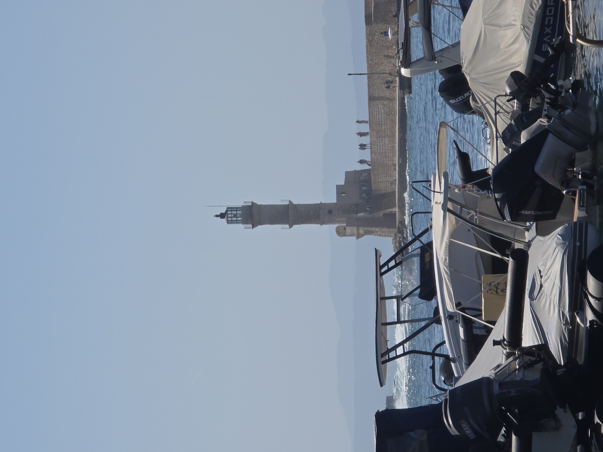 In Chania, Greece, the Venetian Lighthouse stands prominently on a stone quay extending into the Aegean Sea. The lighthouse, constructed from stone, has a cylindrical shape with a smaller lantern room at the top. Several people are visible on the quay, some standing and observing the view, others appearing to walk along the walkway. The sea is active with white-capped waves breaking against the quay. In the foreground, several boats are docked. One boat in the immediate foreground has a dark outboard motor with the brand "YAMAHA" visible. Other boats are covered with tarps, suggesting they are not in use or are being protected. The water is a clear blue, and the sky is a pale, cloudless blue. Distant, hazy mountains can be seen in the background, suggesting a clear but slightly humid day.