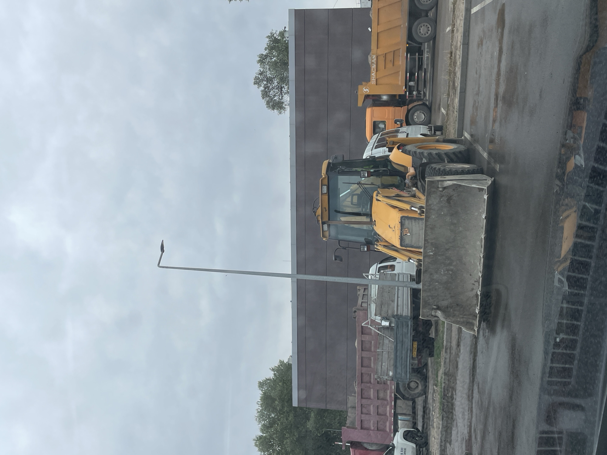 The image shows a cloudy day in Almaty, Kazakhstan, with construction vehicles parked in a lot. A yellow backhoe loader is in the foreground, its shovel resting on the wet asphalt. Behind it are several trucks, including a red dump truck, a white truck with a wooden bed, and an orange SHACMAN dump truck. A modern, dark-brown building with a grey trim runs along the back of the scene, with trees visible to the left and right. A lamppost stands near the center of the frame. The ground is wet, indicating recent rain. There are no people visible in the image.