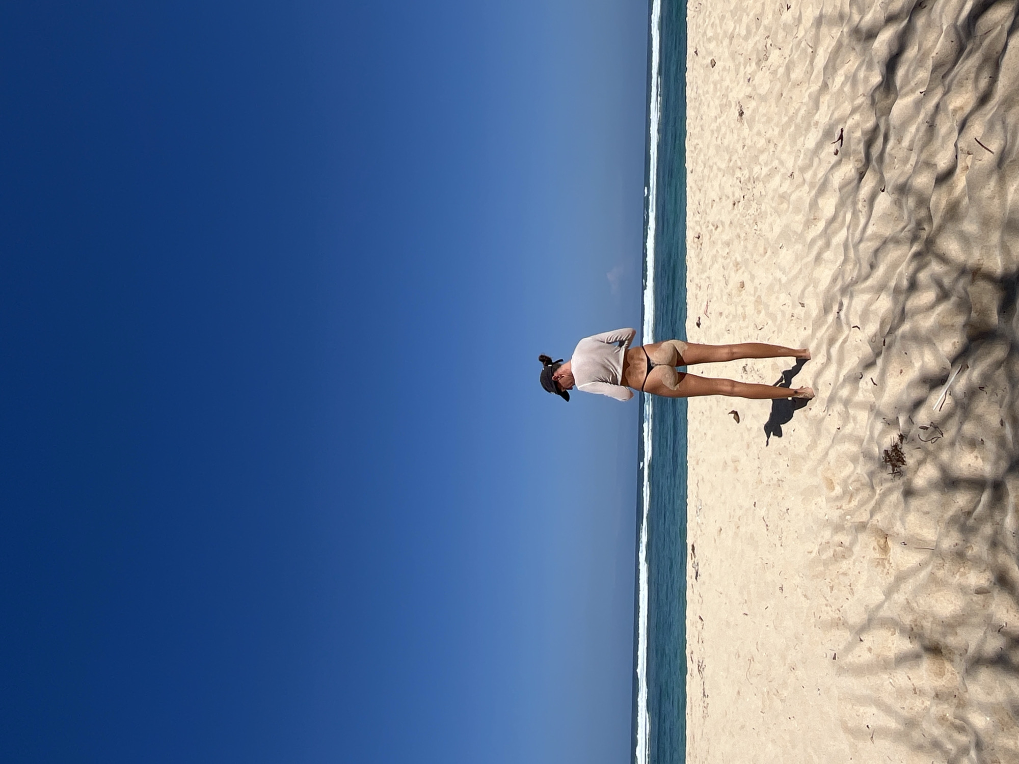 A person stands on a sandy beach in Kuta Selatan, Indonesia, facing away from the camera. They are wearing a light-colored long-sleeved top, a dark baseball cap, and a dark thong bikini bottom. The person appears to be looking down at something in their hands. The beach is wide and covered in light-colored sand, with some scattered debris and shadows cast by unseen objects. The ocean stretches out to the horizon, with small waves breaking near the shore. The sky is a clear, deep blue.