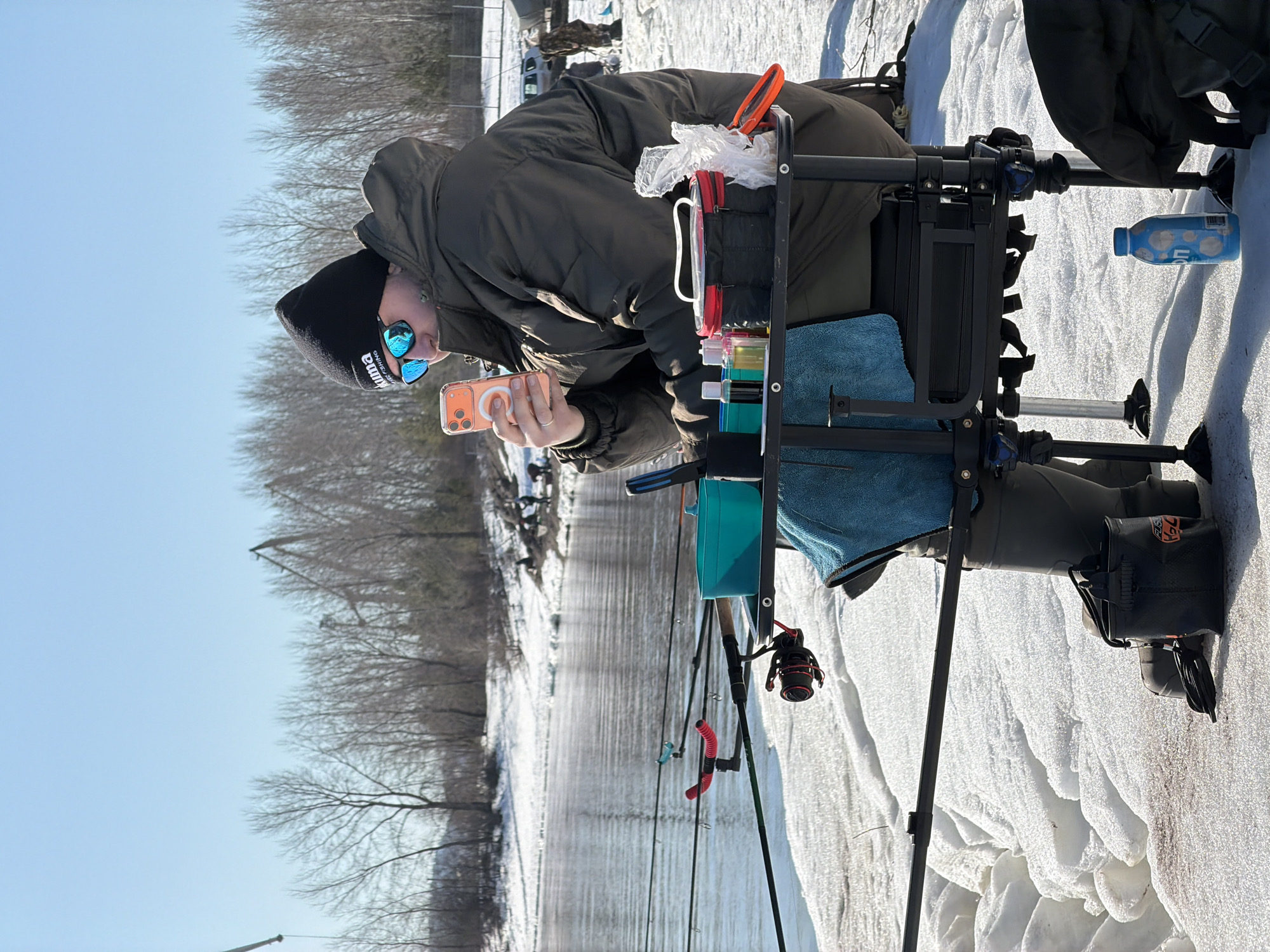 A person in a black beanie with white lettering, reflective blue sunglasses, and a dark green puffy coat is sitting on a portable fishing seat. The person is holding a peach-colored smartphone, appearing to be taking a photo or interacting with the screen. The fishing seat is equipped with containers, a towel, and other fishing gear. In the background, a river runs along the edge of a snowy landscape, flanked by bare trees and buildings. The scene suggests an outdoor winter fishing setting. While the description included a location in the city of Noviy, Russia, it is not definitively determined from the image alone.